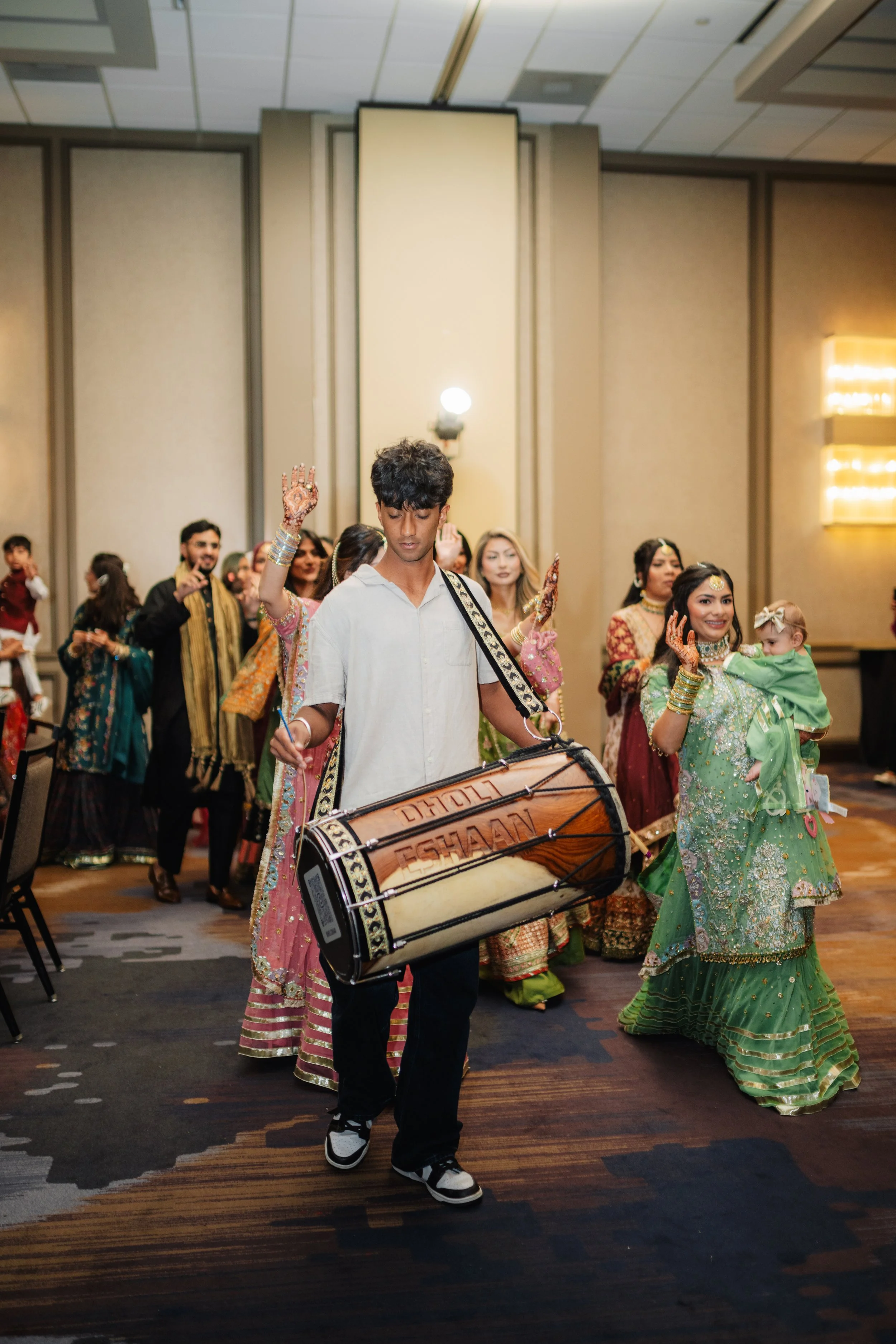 People dressed in traditional Indian clothing participating in a cultural dance, with a person in the foreground playing a large drum, in a banquet hall setting.