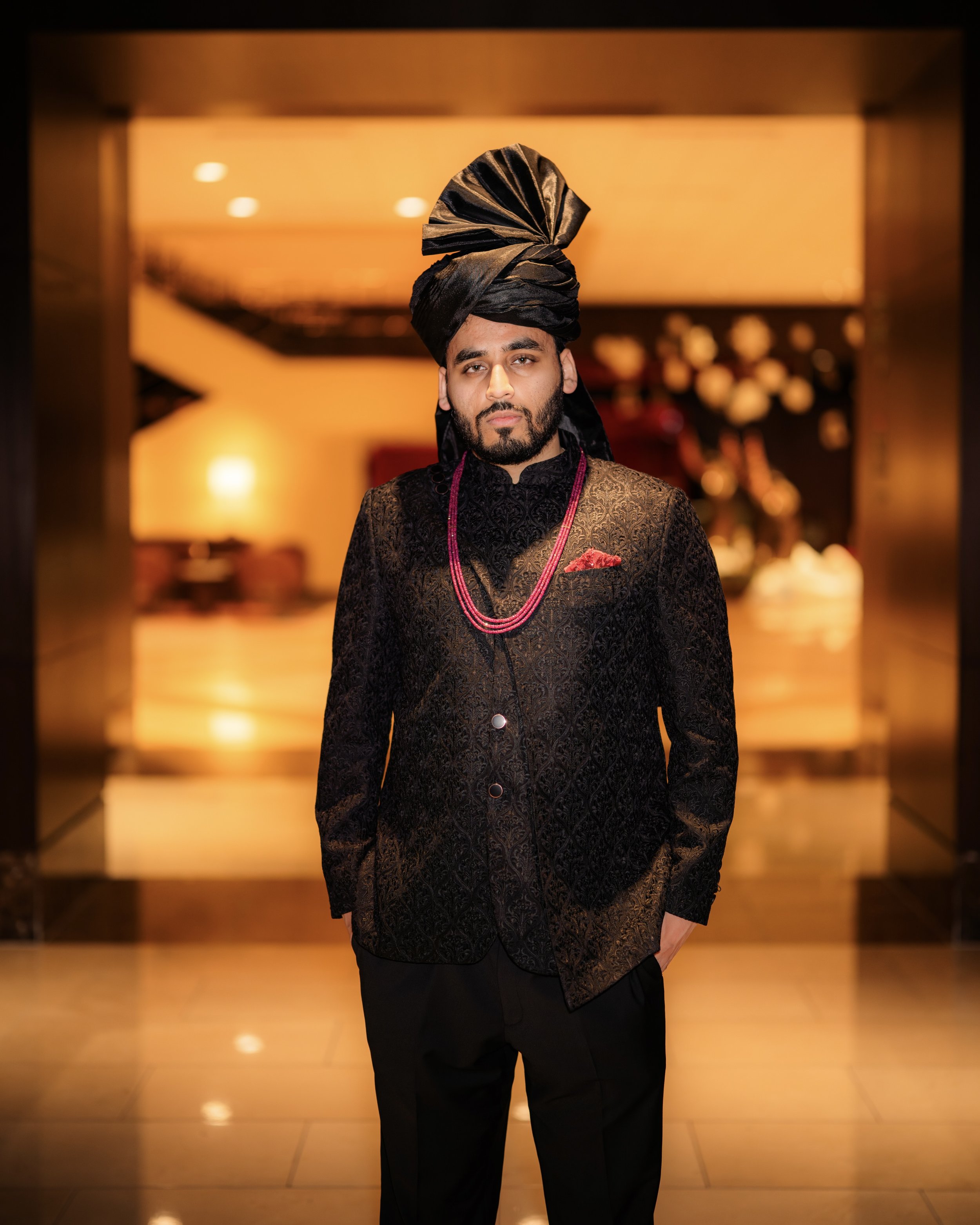 Man dressed in traditional Indian attire with a turban and jewelry, standing indoors with warm lighting.