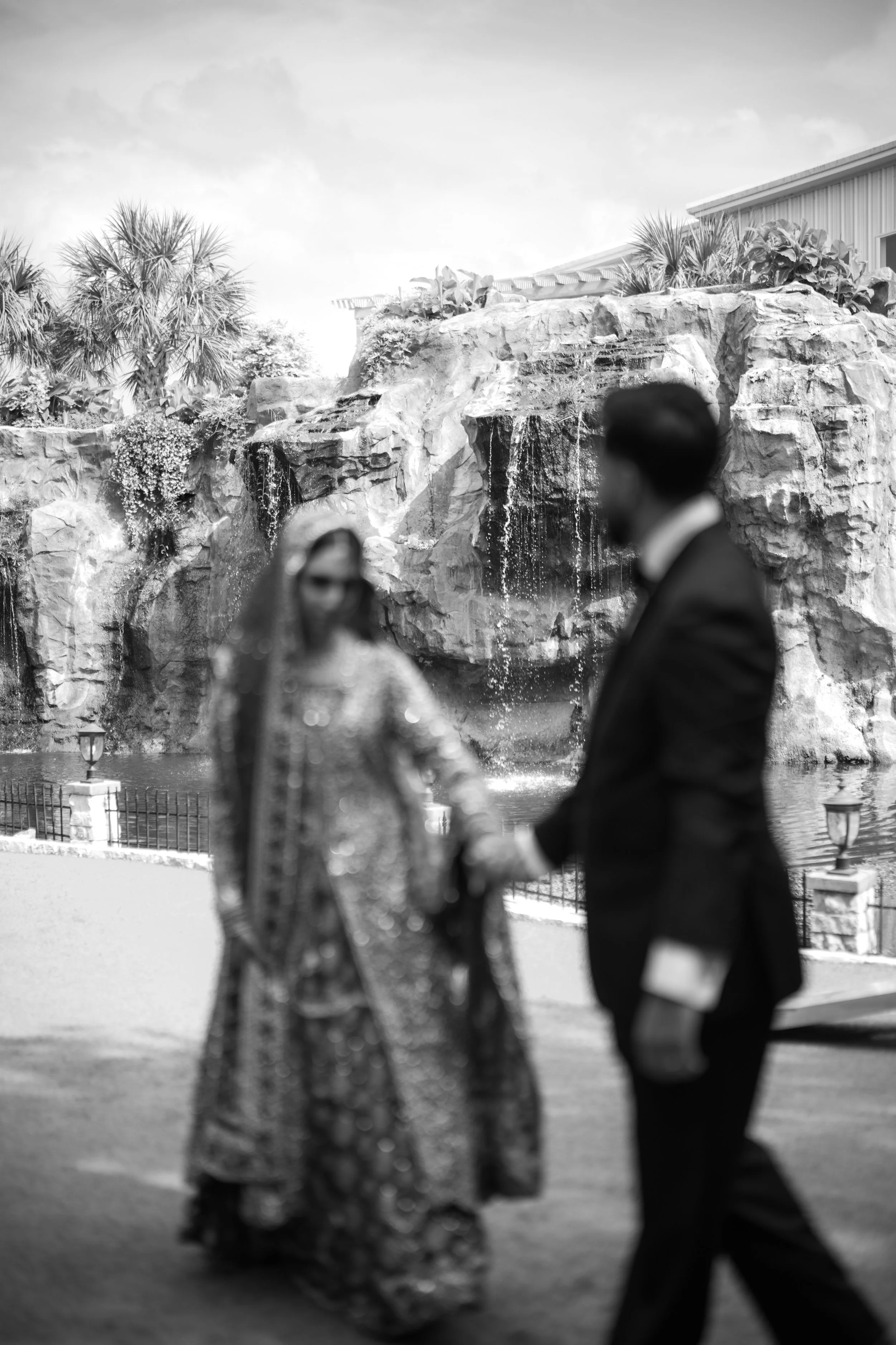 A man and woman holding hands, dressed in formal attire, standing outdoors near a waterfall and lush tropical plants.