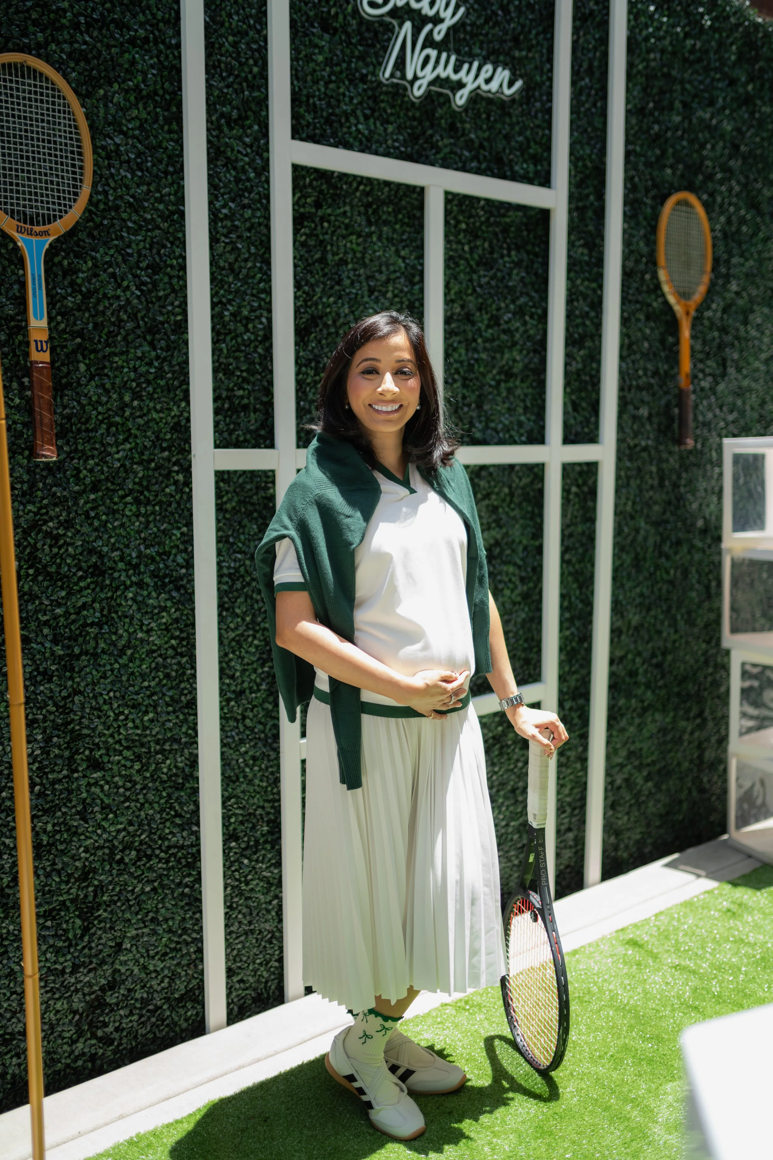 A woman standing on artificial grass with a tennis racket, wearing a white and green sports outfit, sunglasses, and sneakers, in front of a green hedge wall with white framing and a neon sign that says 'Lucky Nguyen'.