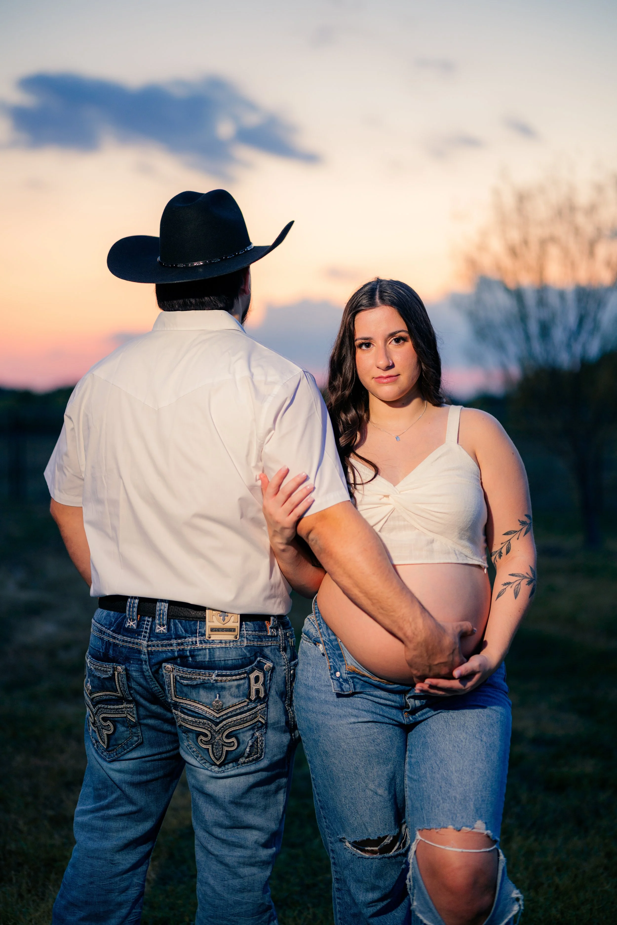 A pregnant woman with dark hair and a tattoo on her arm stands outdoors at sunset, gently holding her belly. A man in a white shirt and black cowboy hat is standing close to her, with one hand on her belly and the other arm around her back. The backg
