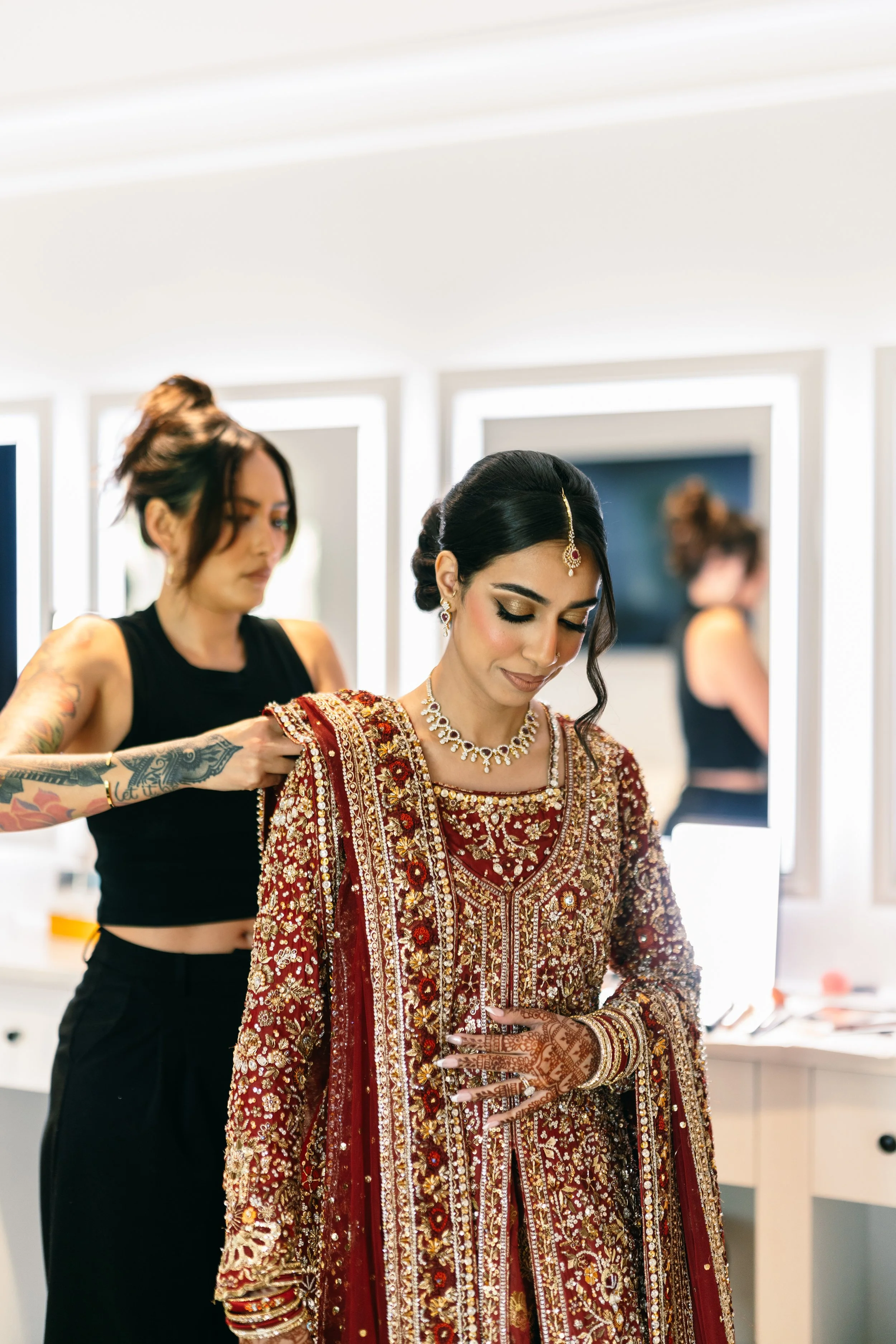 A bride dressed in a traditional red and gold embroidered bridal gown, adorned with jewelry, madly looking at her phone. A stylist assists her with her outfit, both standing in front of mirrors in a well-lit dressing room.