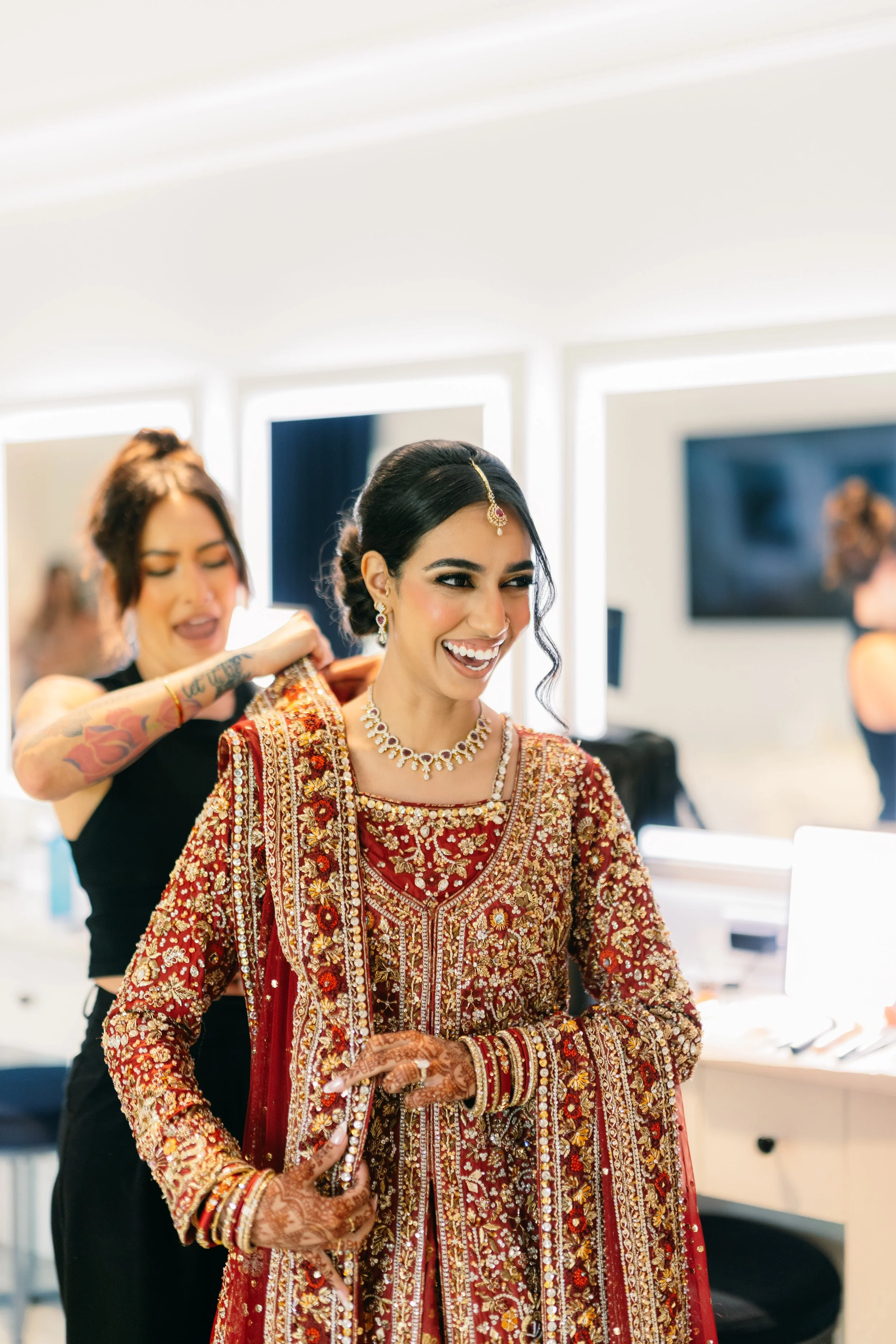 A bride dressed in traditional South Asian wedding attire, wearing an ornate red and gold embroidered dress, jewelry including necklace, earrings, and forehead ornament, with henna on her hands, smiling.