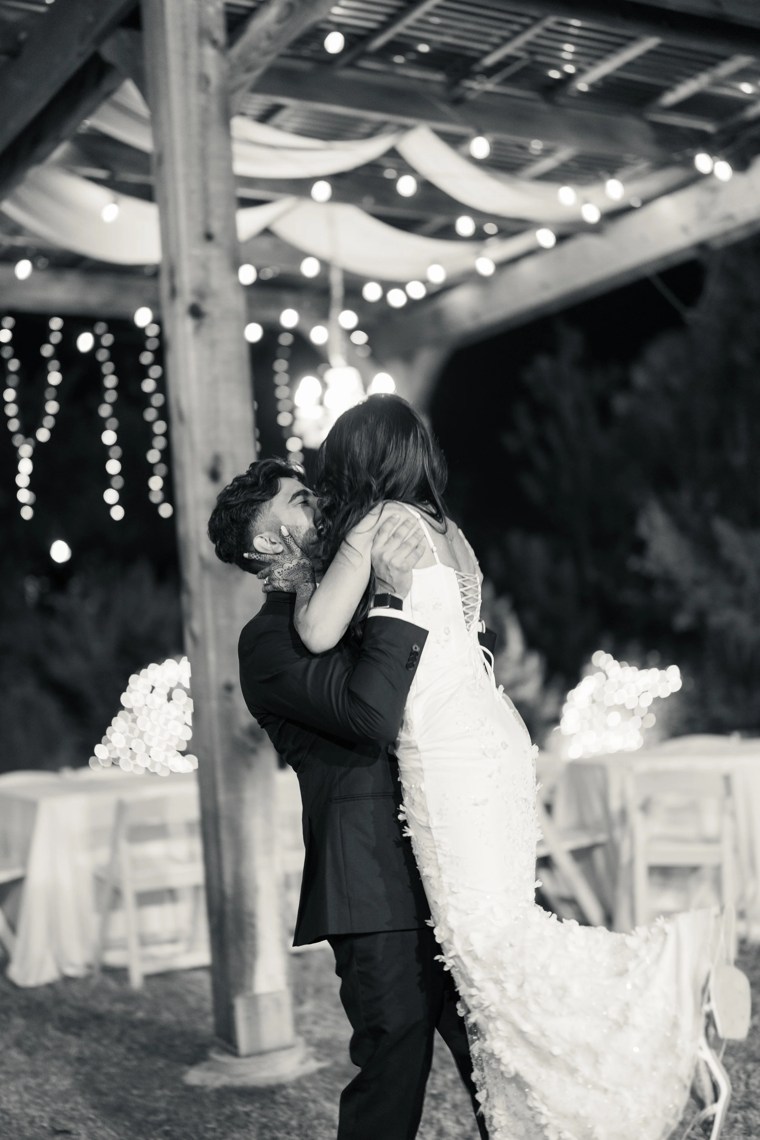 A man in a black suit lifts a woman in a white wedding dress during a wedding celebration at night, with string lights and a decorated outdoor setting.