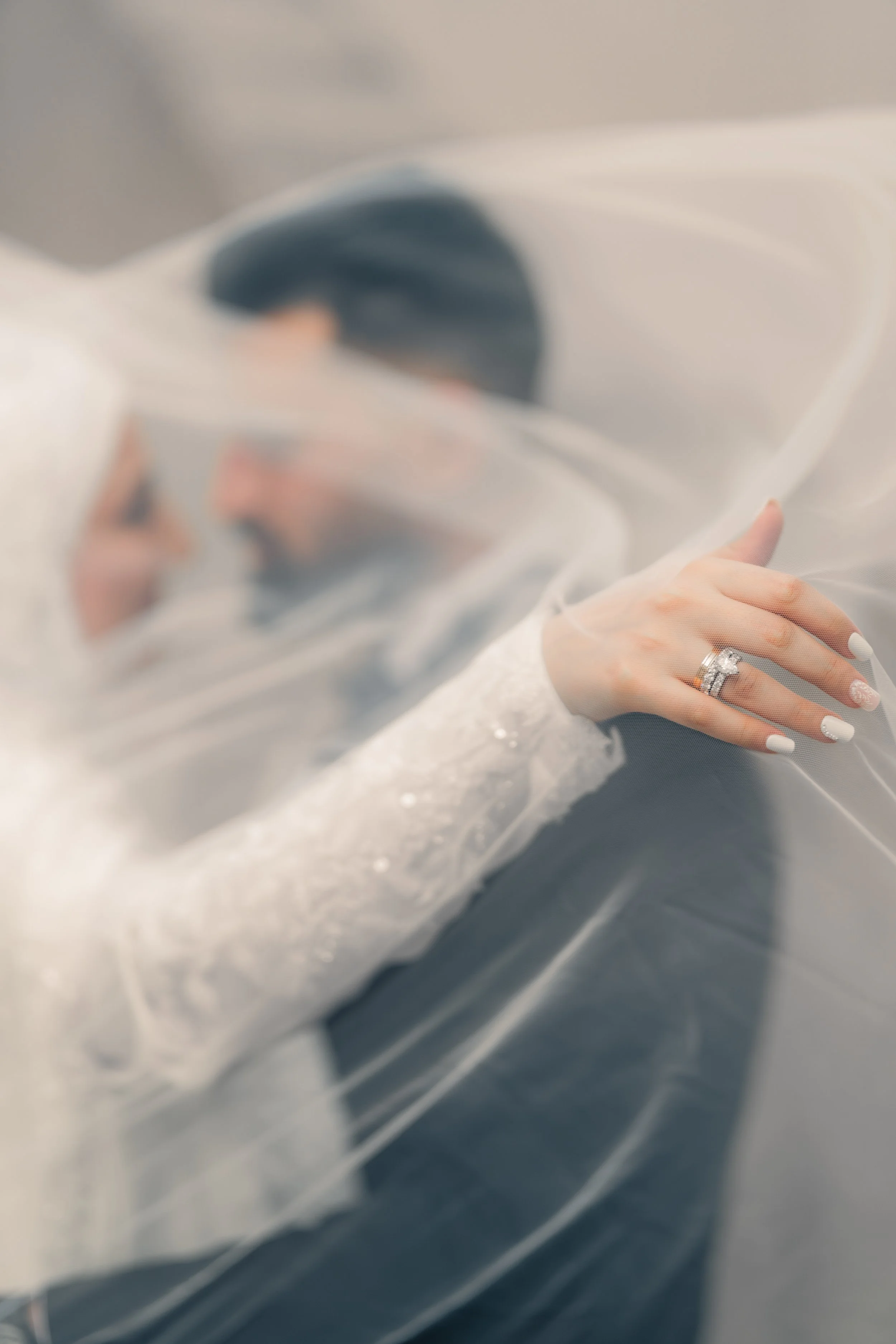 Close-up of a bride's hand with wedding rings, resting on her gown, covered by a sheer veil. The background shows a blurred image of the bride and groom leaning close together.