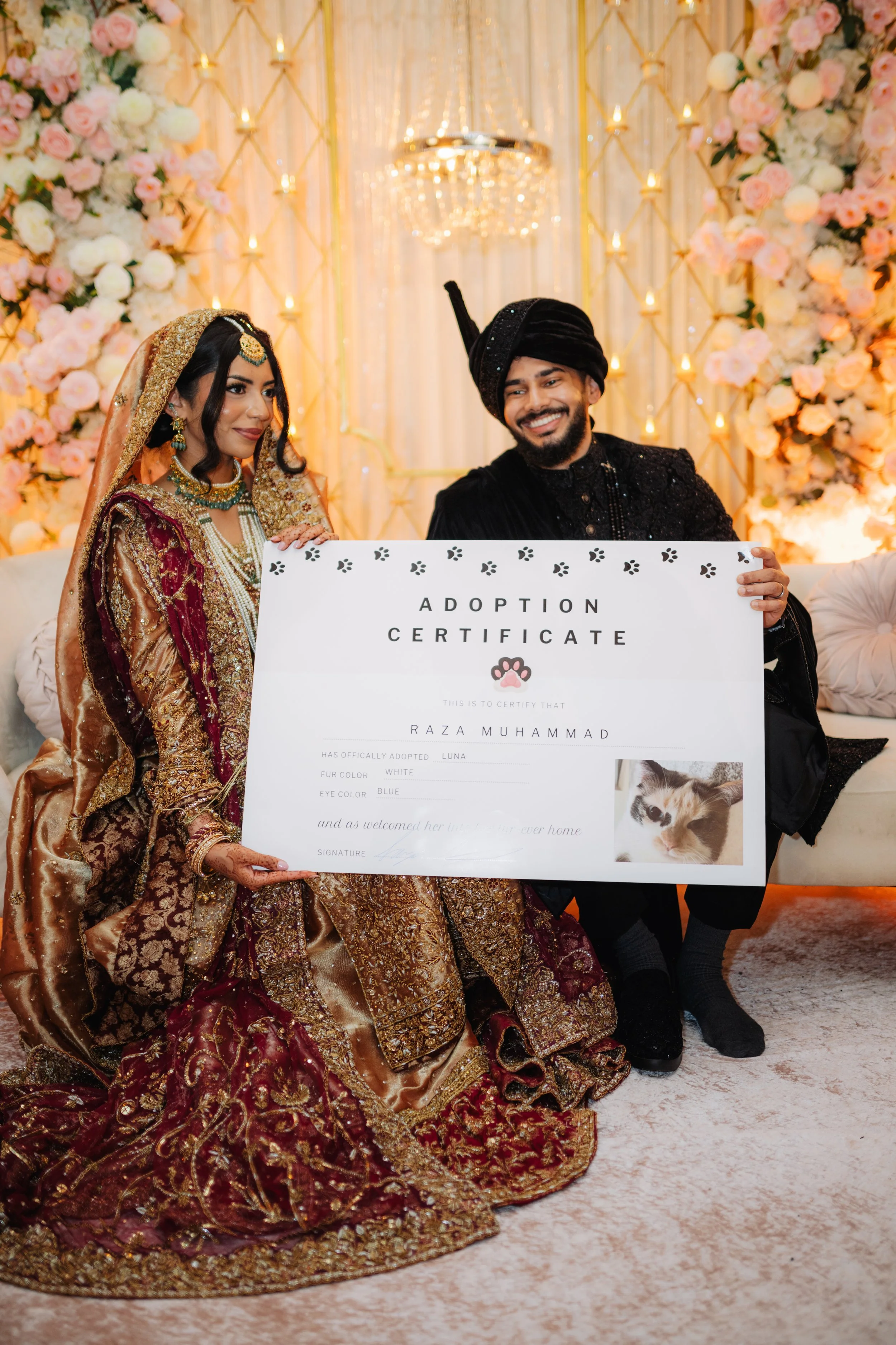 A woman in traditional South Asian bridal attire sitting on the floor, holding an adoption certificate with a photo of a cat, while a man in traditional attire standing beside her holds the certificate as well, in a decorated indoor setting with flor