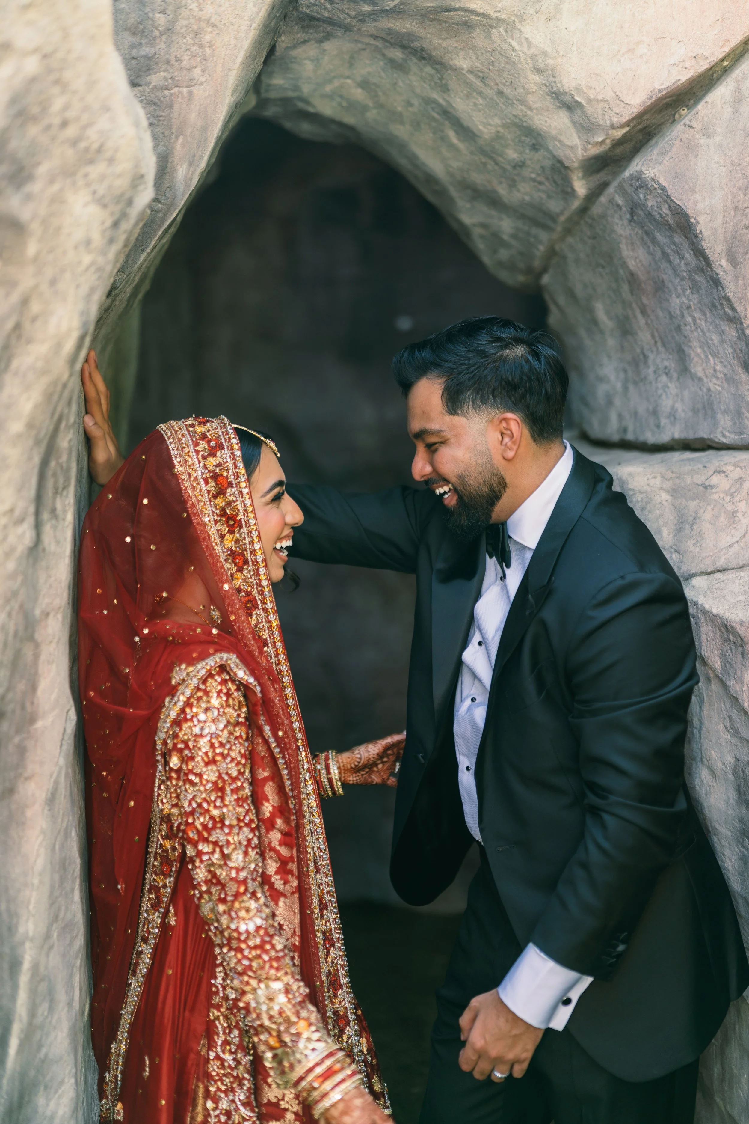 A couple of Indian descent in traditional wedding attire inside a stone alcove. The woman is dressed in a red embroidered bridal outfit with a headscarf, and the man is in a black tuxedo. They are smiling and holding hands, with the man leaning towar