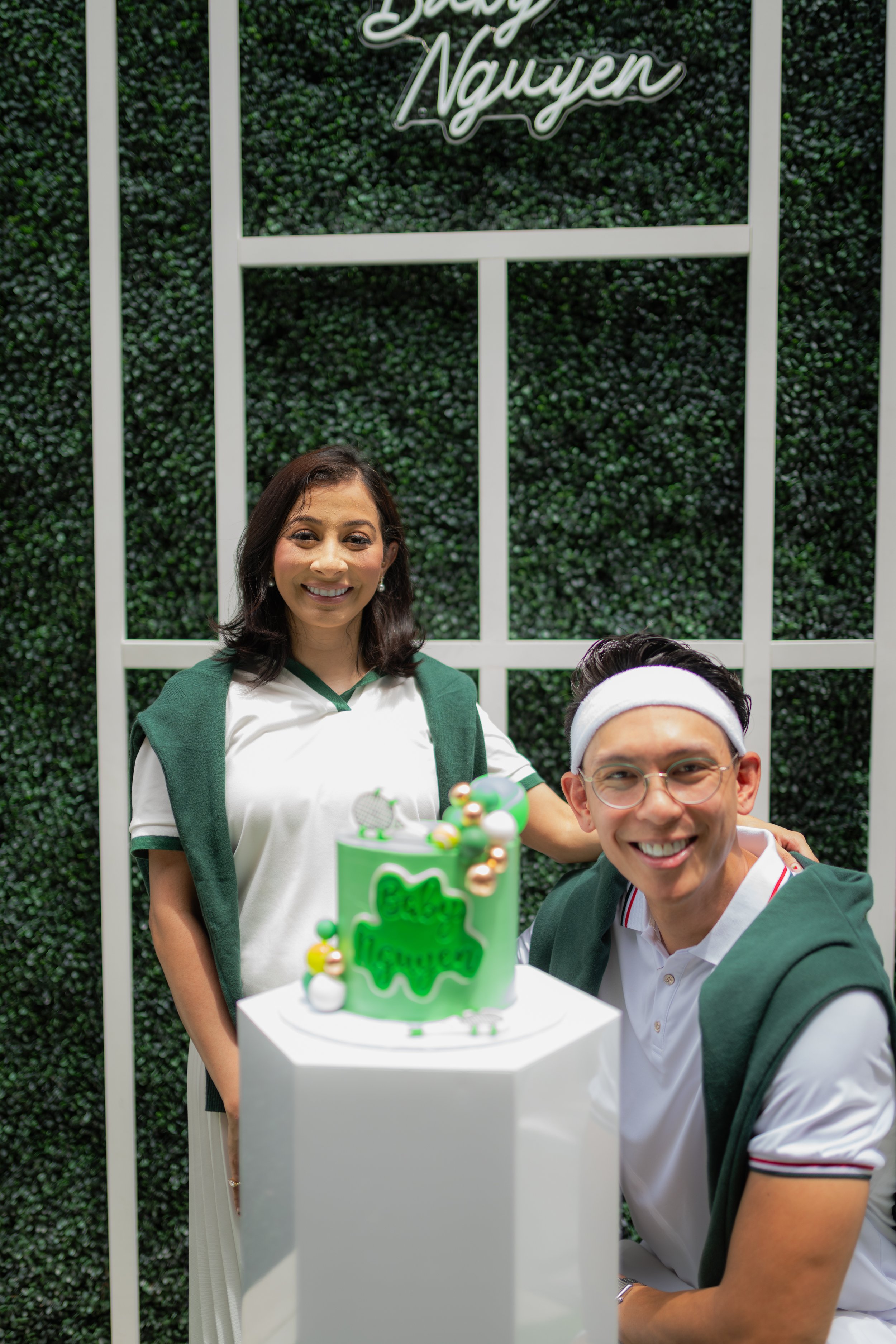Two people celebrate a birthday in front of a green leafy background with a sign that reads "Birthday Nayen." The woman on the left is smiling and wearing a green and white shirt. The man on the right is also smiling, wearing glasses and a white shir