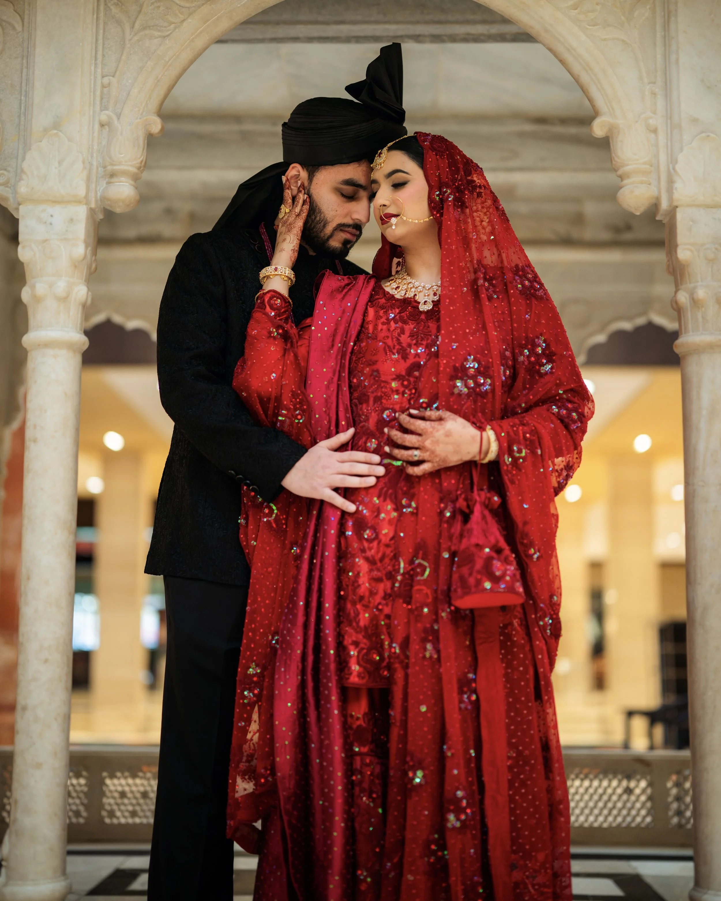 A couple in traditional Indian wedding attire standing close together under an ornate archway in an indoor setting. The bride wears a red saree with intricate embroidery and jewelry, and the groom wears a black sherwani with a matching turban.