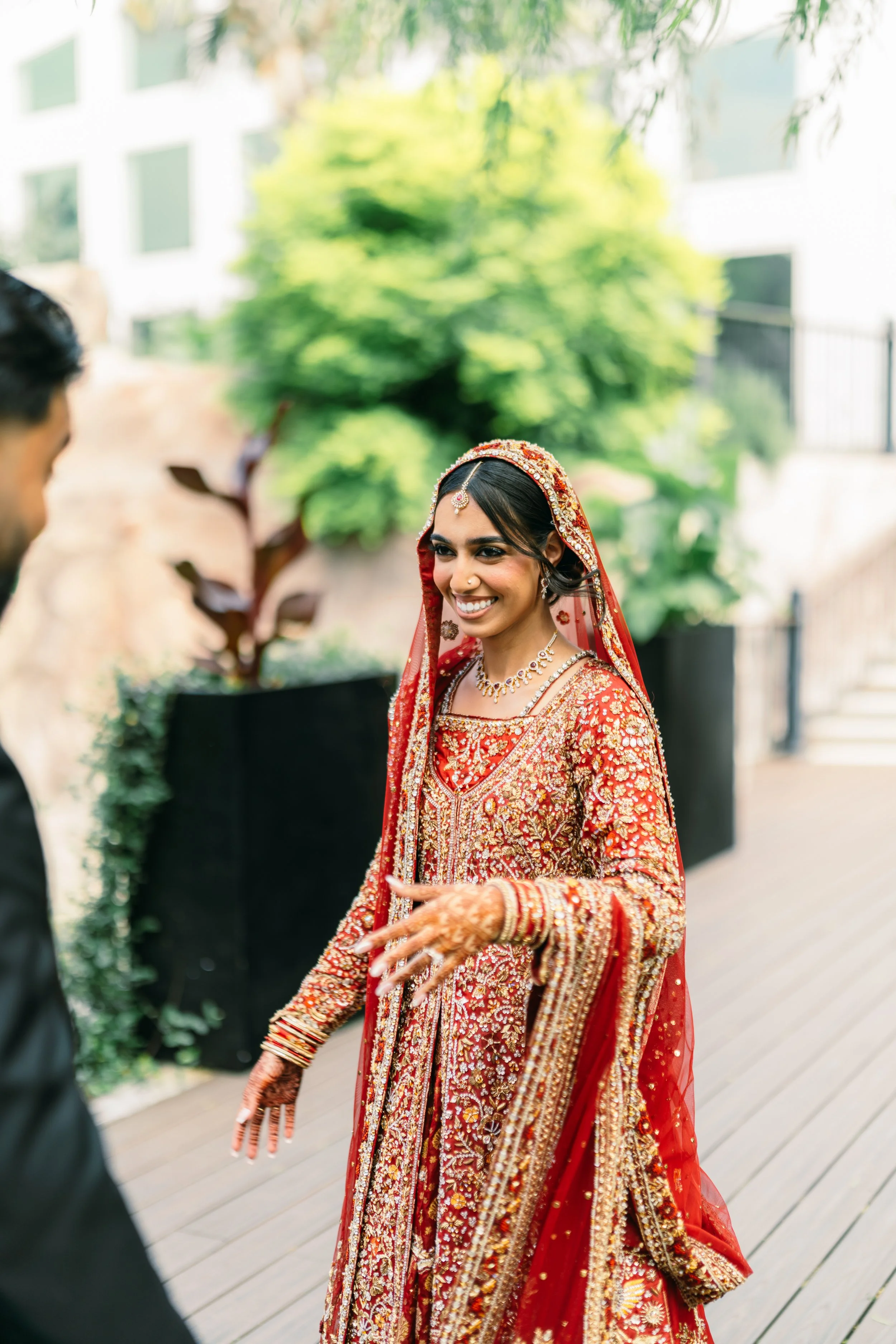 A woman dressed in traditional red and gold wedding attire, smiling and holding hands with a person in a black suit, outdoors with greenery in the background.