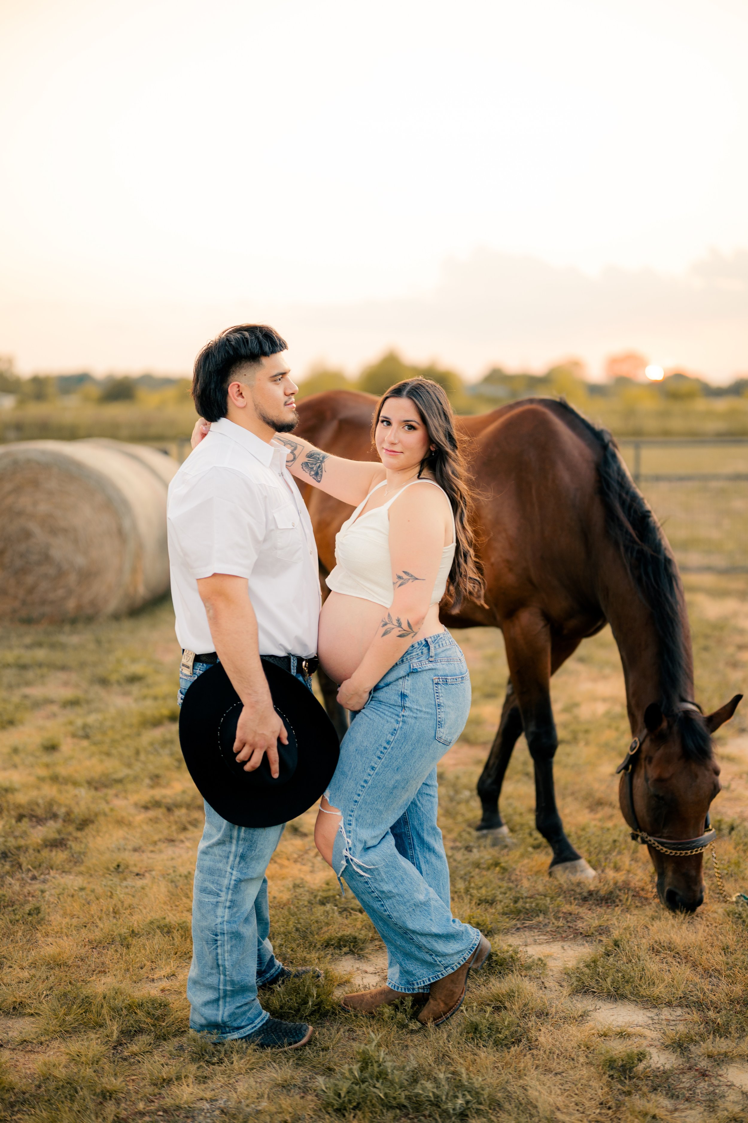 A pregnant woman and a man standing together outdoors with a horse in the background. The woman has tattoos and is holding her belly, wearing a cream crop top and ripped jeans. The man, holding a black hat, is wearing a white shirt and jeans. The set
