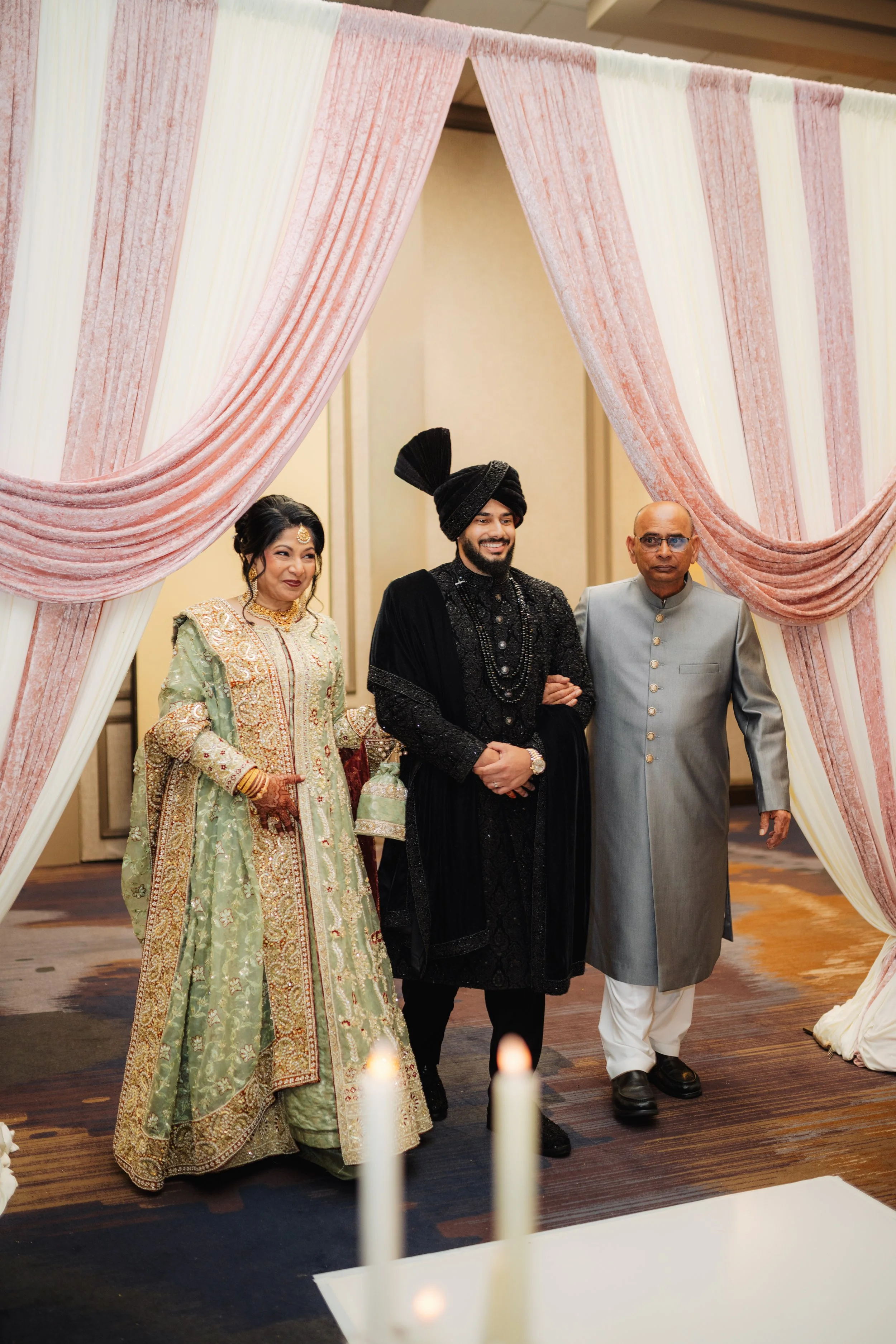 Three people at a wedding celebration standing under pink and cream draped fabric decor. The woman is dressed in a light green traditional outfit with gold and red embroidery, the man in a black traditional Indian outfit with a black turban, and the 