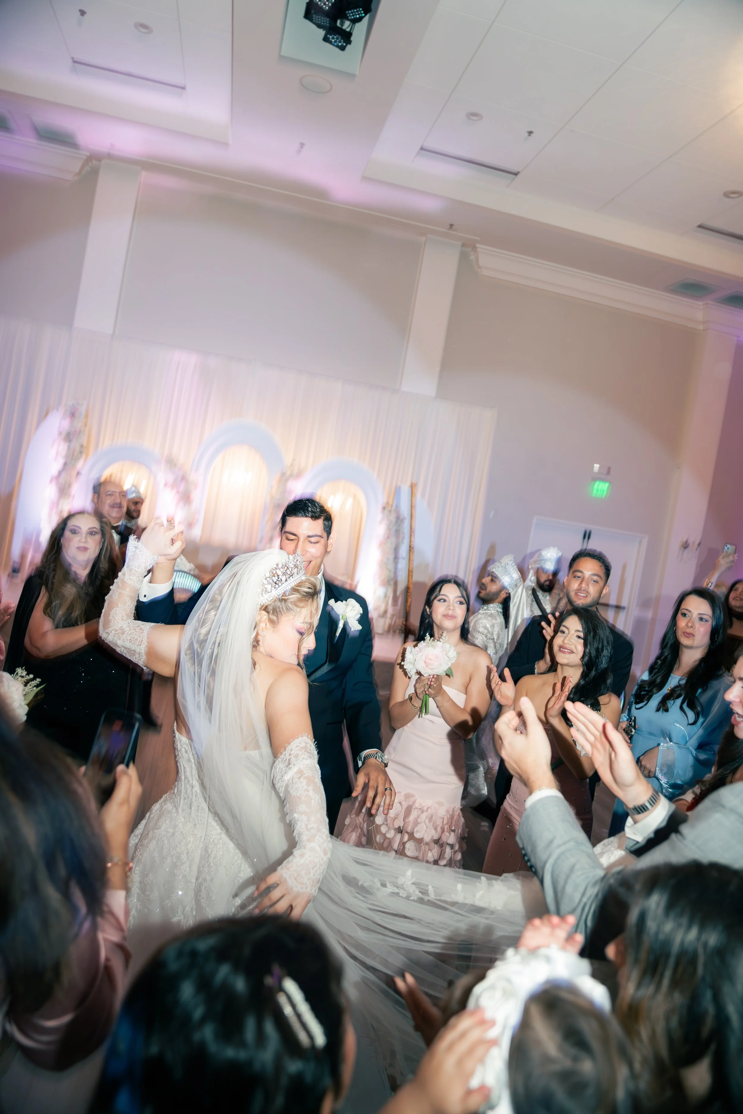 A bride and groom dancing at their wedding reception surrounded by friends and family applauding and celebrating.