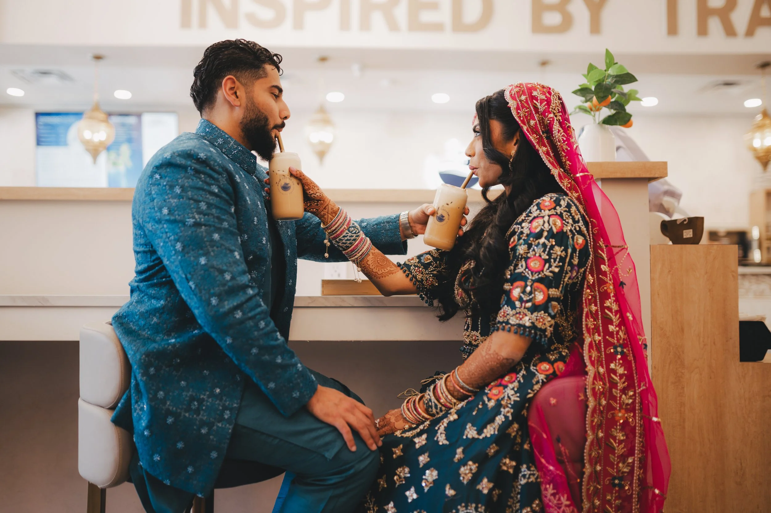 A woman dressed in traditional South Asian wedding attire is sharing a drink with a man dressed in a blue suit in a modern indoor setting, likely a wedding or celebration.