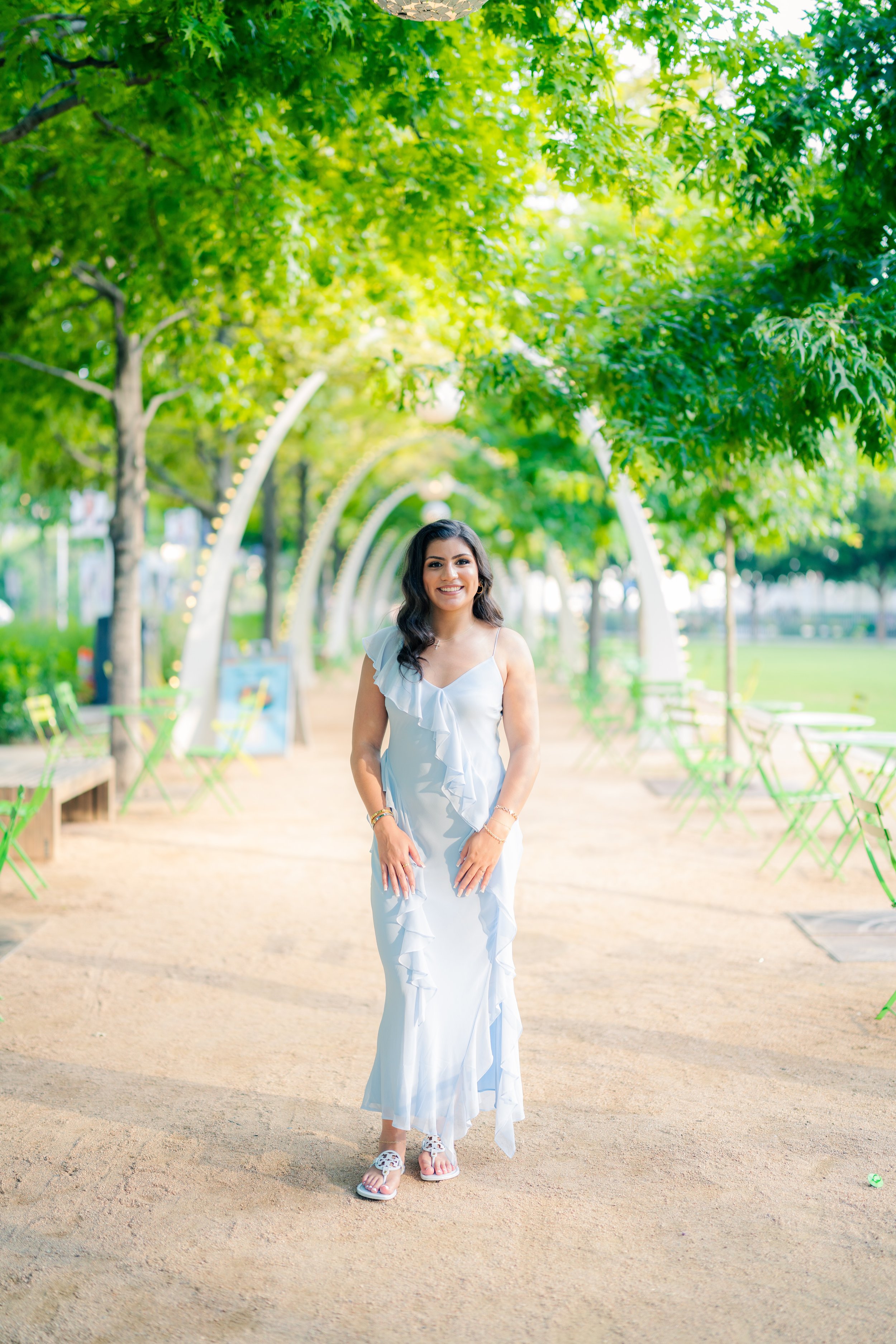 Smiling woman in white dress standing on a dirt path in a park with green trees and chairs.