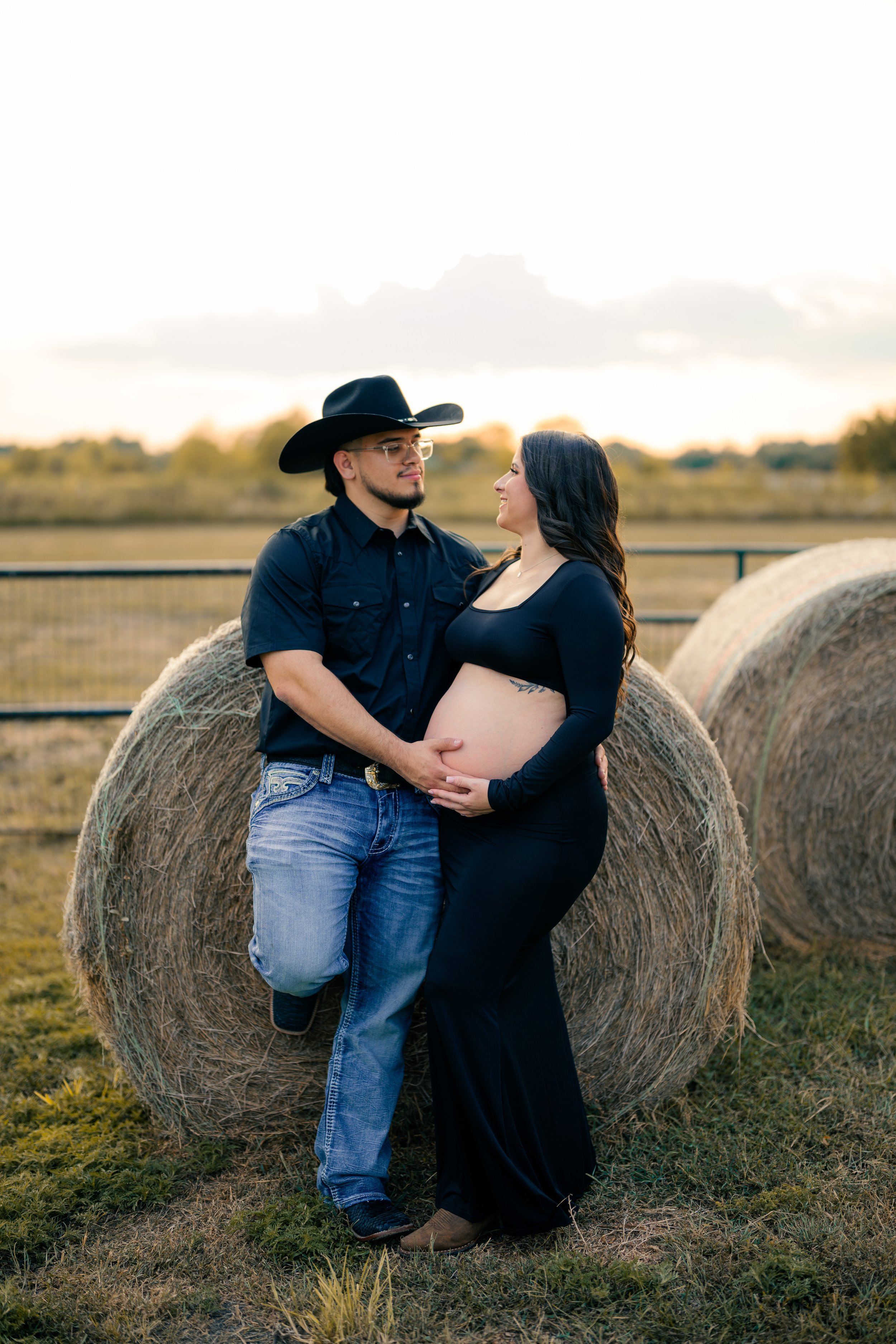 A pregnant woman and man standing next to hay bales in an outdoor farm setting during sunset, holding her belly and looking at each other.