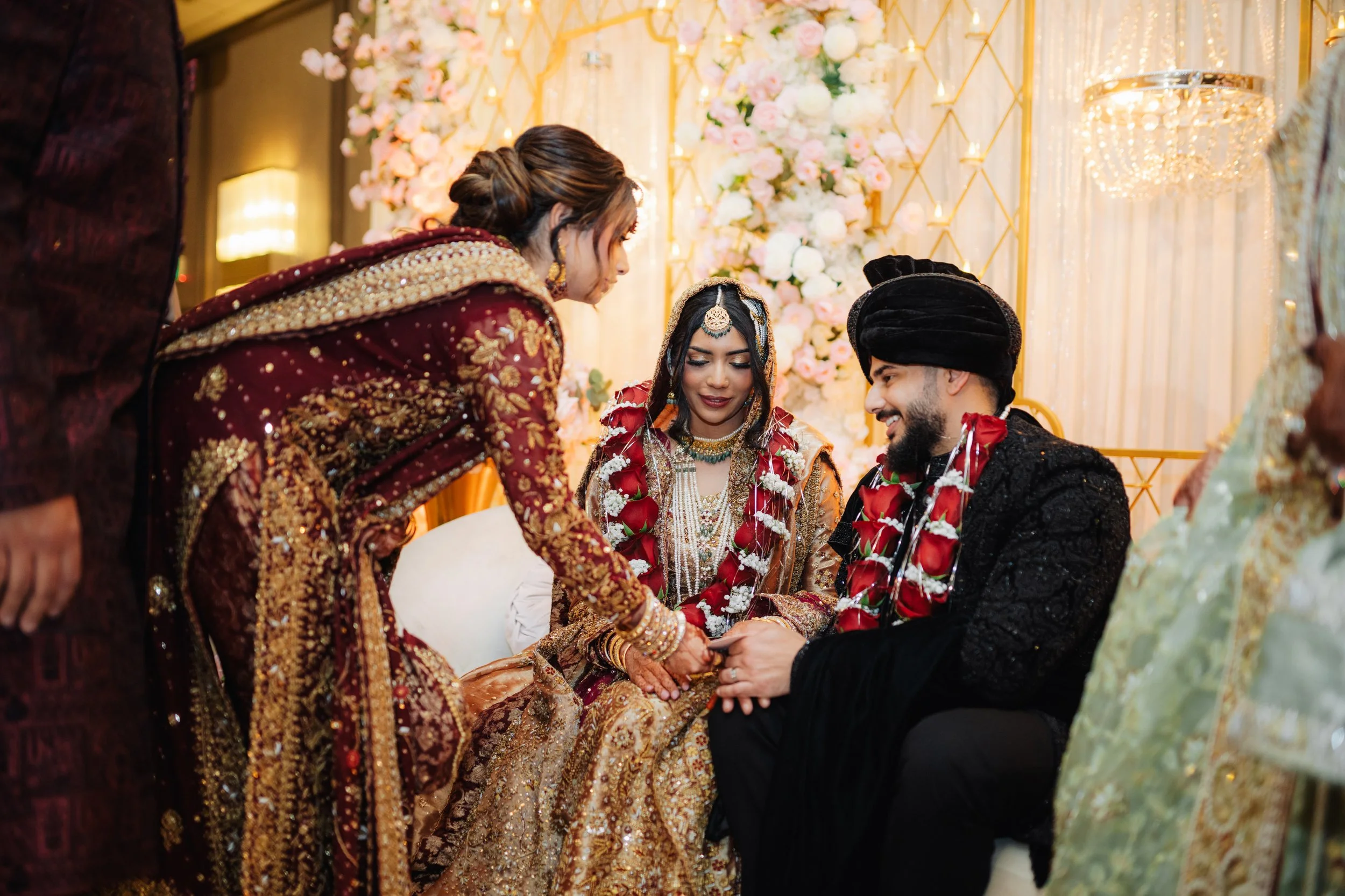 Indian bride and groom during wedding ceremony; woman in maroon and gold traditional attire, man in black traditional outfit, surrounded by family and floral decorations.