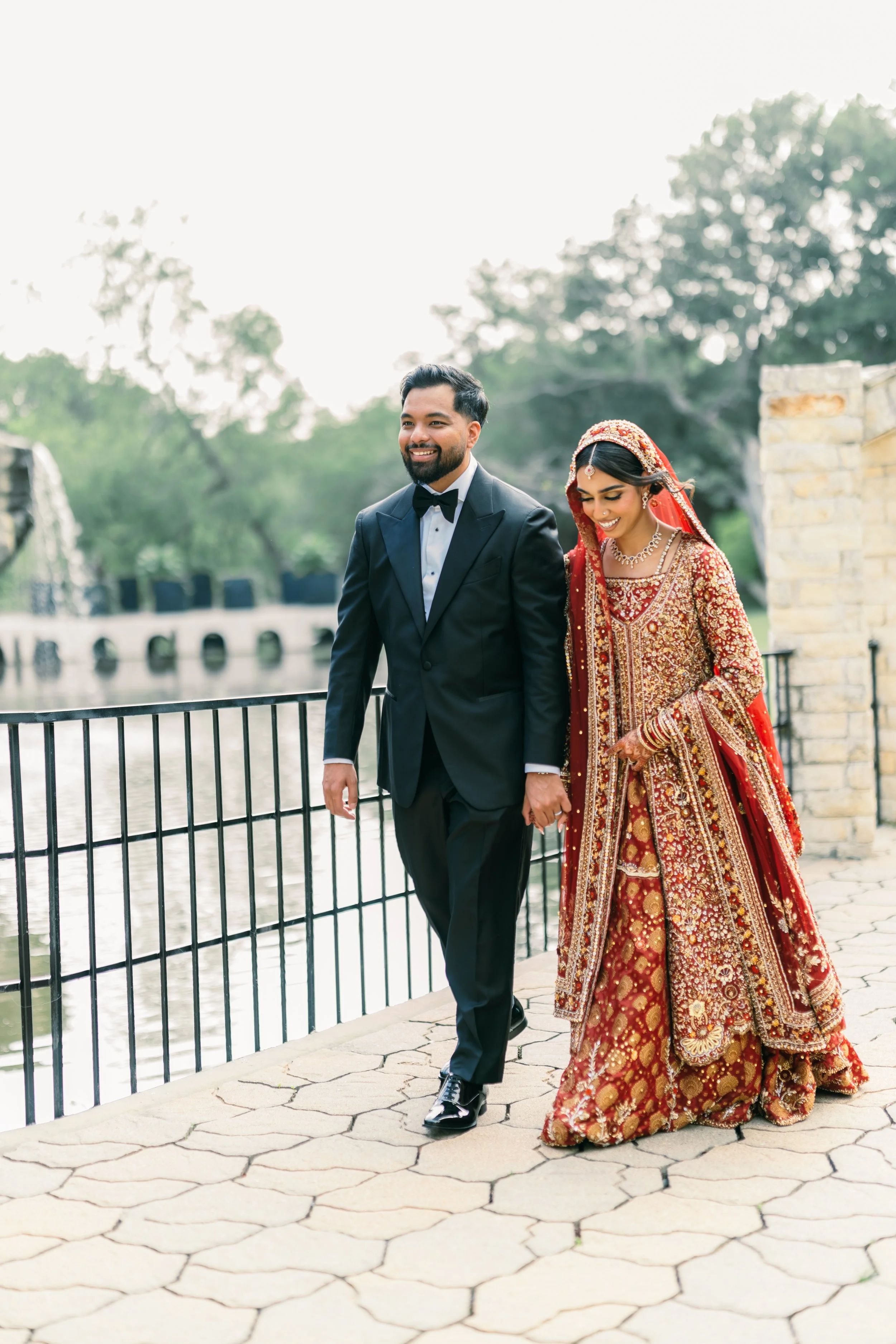 A man in a tuxedo and a woman in a traditional red and gold Indian wedding dress walking near a body of water with trees in the background.