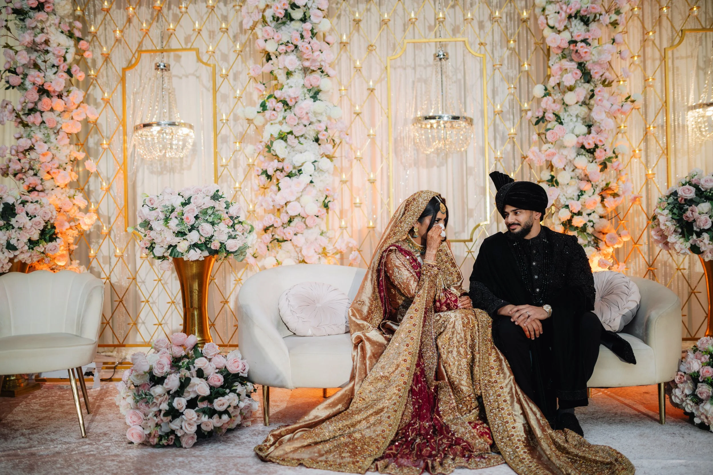 A bride and groom seated on a cream-colored sofa at a wedding reception, surrounded by pink and white floral arrangements and chandeliers.