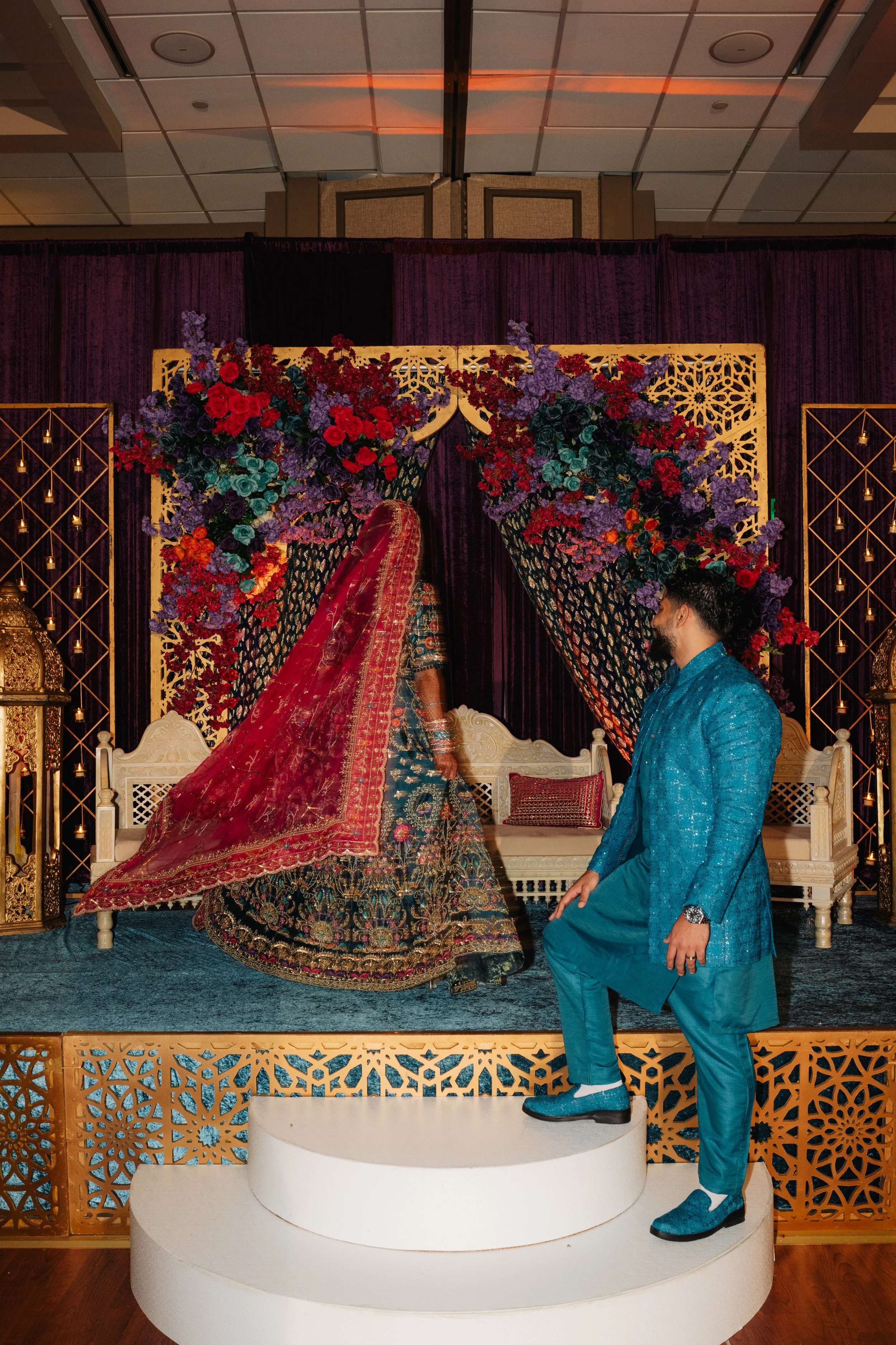 A woman in traditional Indian attire is standing on a white platform, facing a man who is kneeling before her in an elaborately decorated indoor setting with flowers and ornate panels.