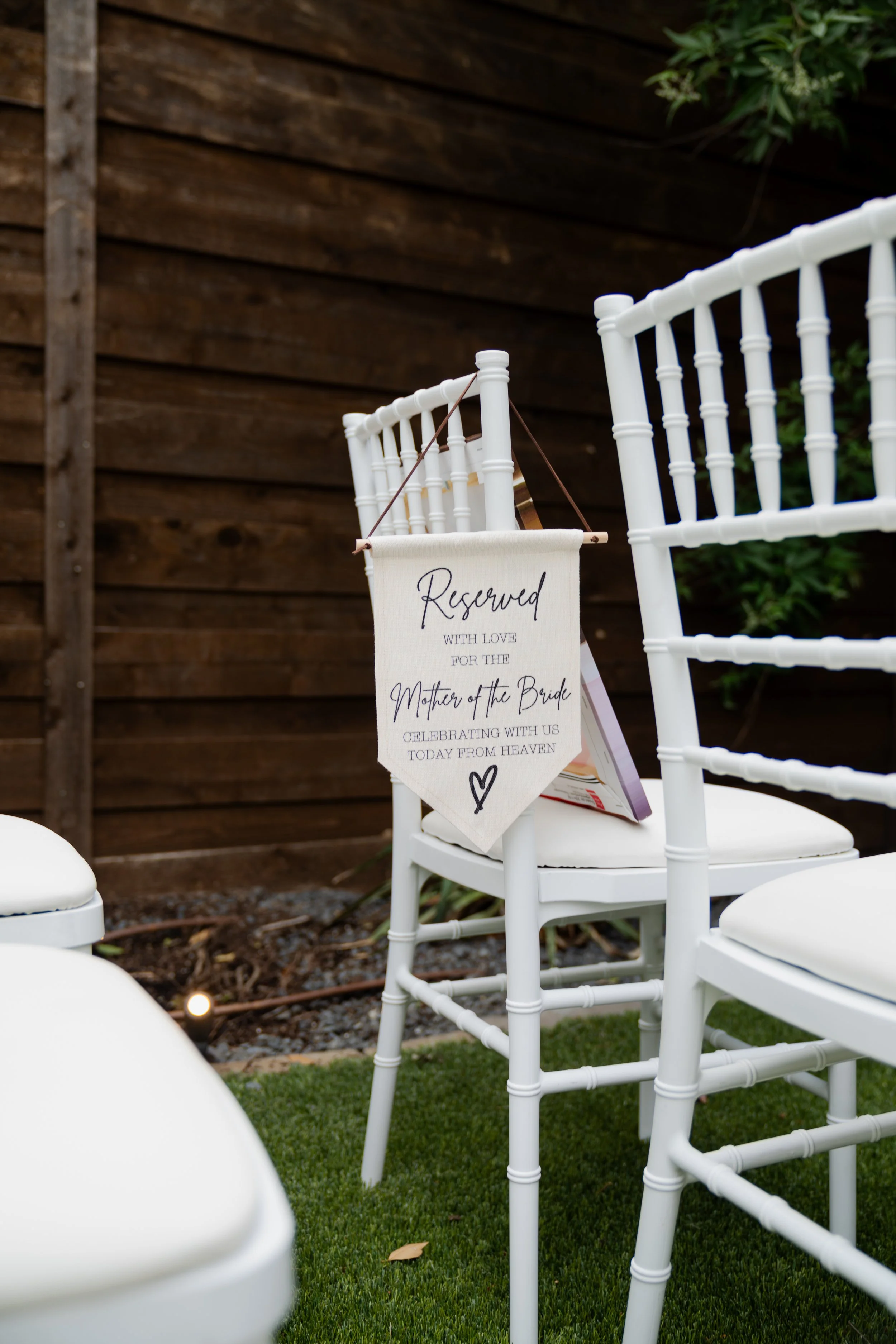 White chairs are arranged outdoors on a green lawn with a wooden fence and garden plants in the background. One chair has a small fabric flag hanging from it that says, 'Reserved with love for the Mother of the Bride, celebrating with us today from h