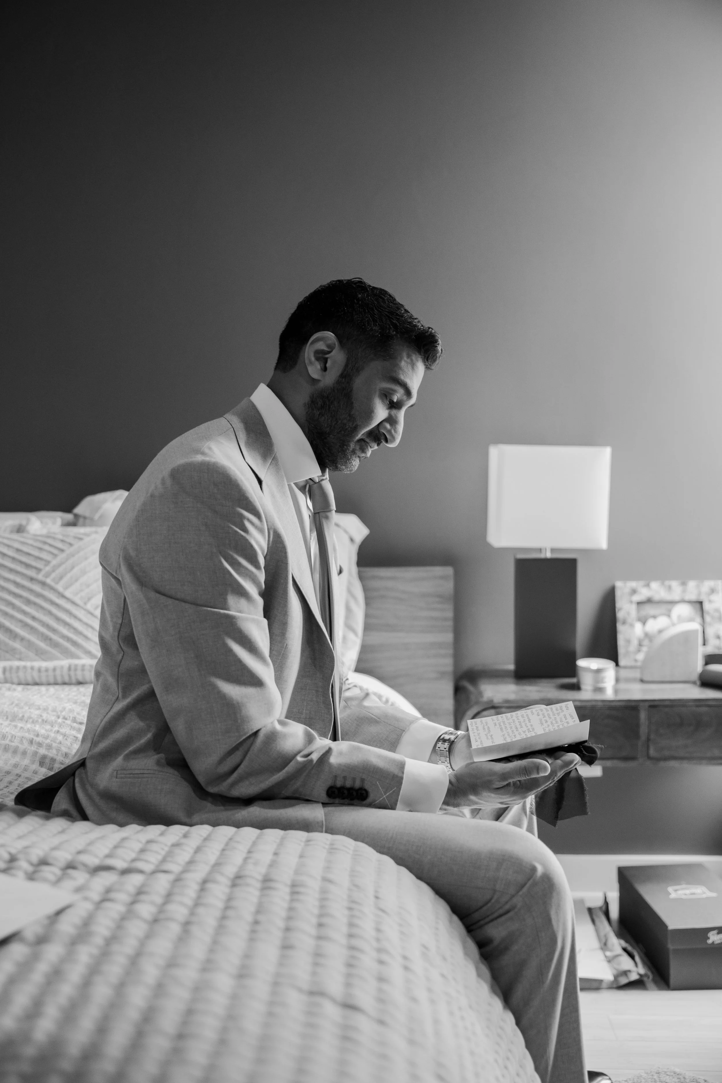 A man in a suit sitting on a bed, reading a book in a dimly lit room with a bedside lamp.