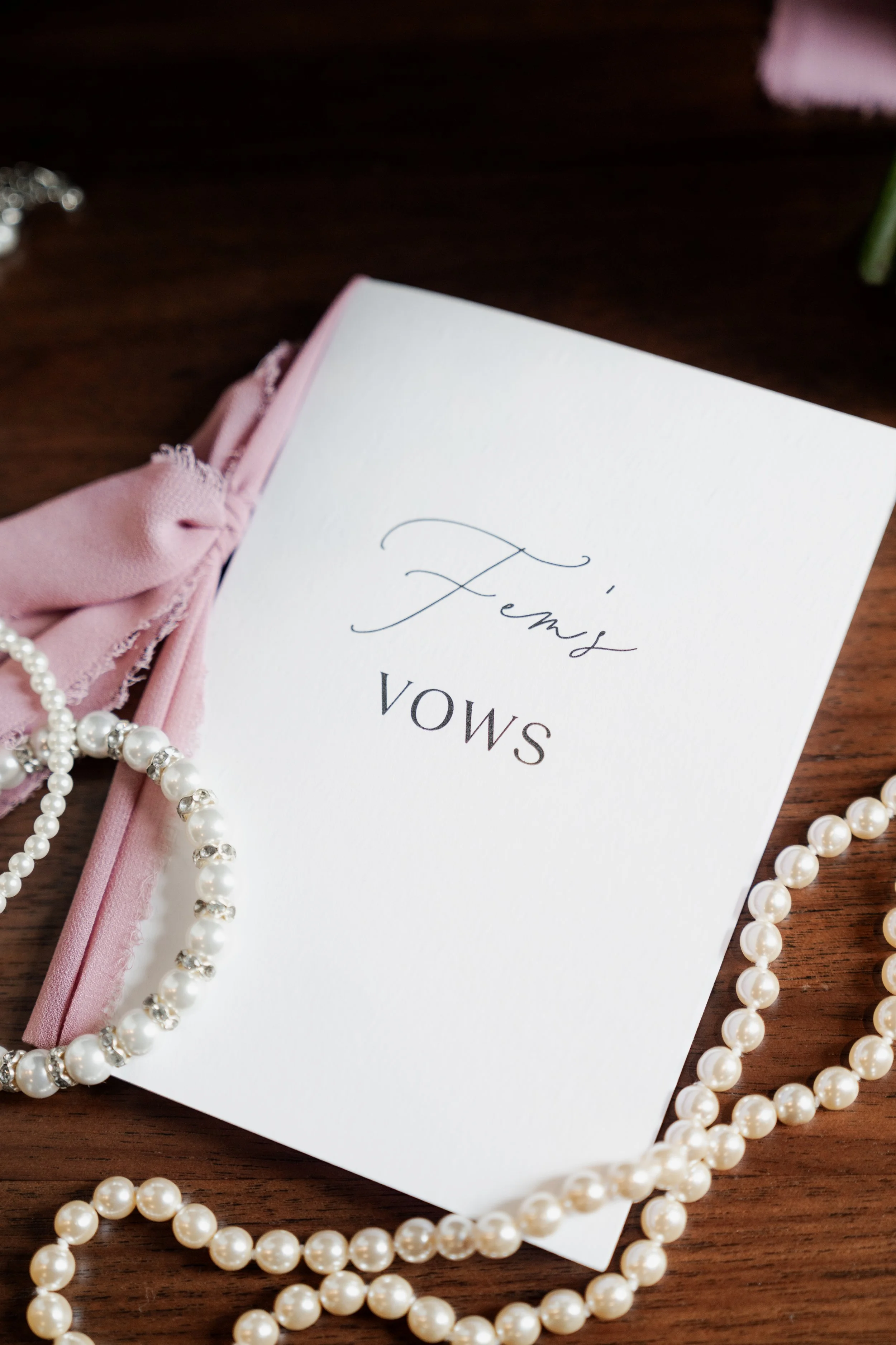 A wedding vow booklet with the word 'Femi's' written on the cover, placed on a wooden surface. There are pearls both above and below the booklet, and a pink cloth with fraying edges partially wrapped around the booklet.