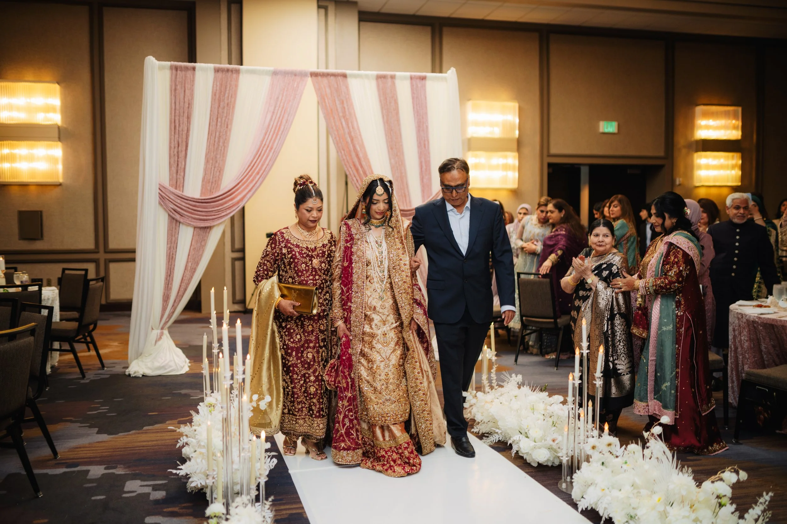 A bride and groom in traditional wedding attire walking down an aisle decorated with white flowers and candles, attended by family and guests at a wedding reception.