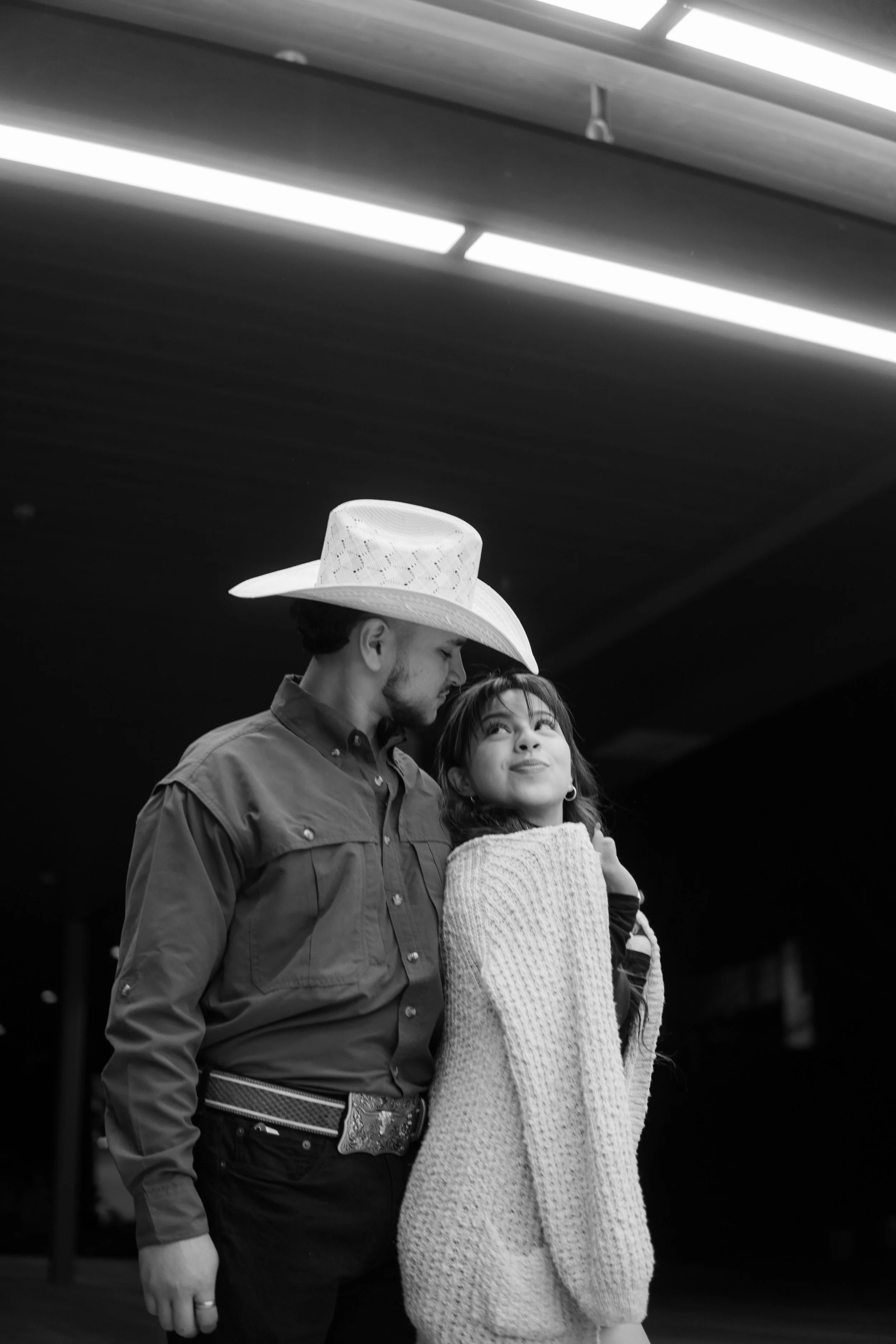 A black and white photo of a man wearing a cowboy hat and a woman wrapped in a blanket, standing close together indoors under a ceiling with fluorescent lights.