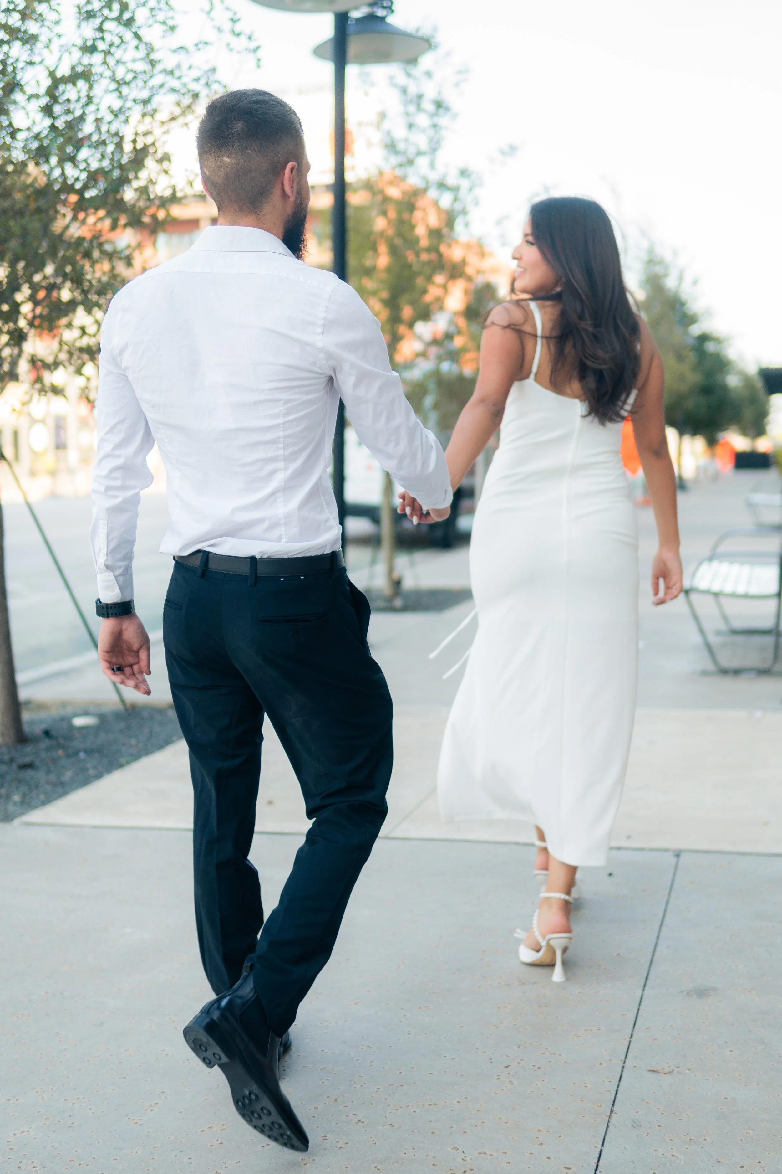 A couple holding hands and walking outside, dressed in formal attire. The man is wearing a white shirt and black pants, and the woman is wearing a white dress and high heels.