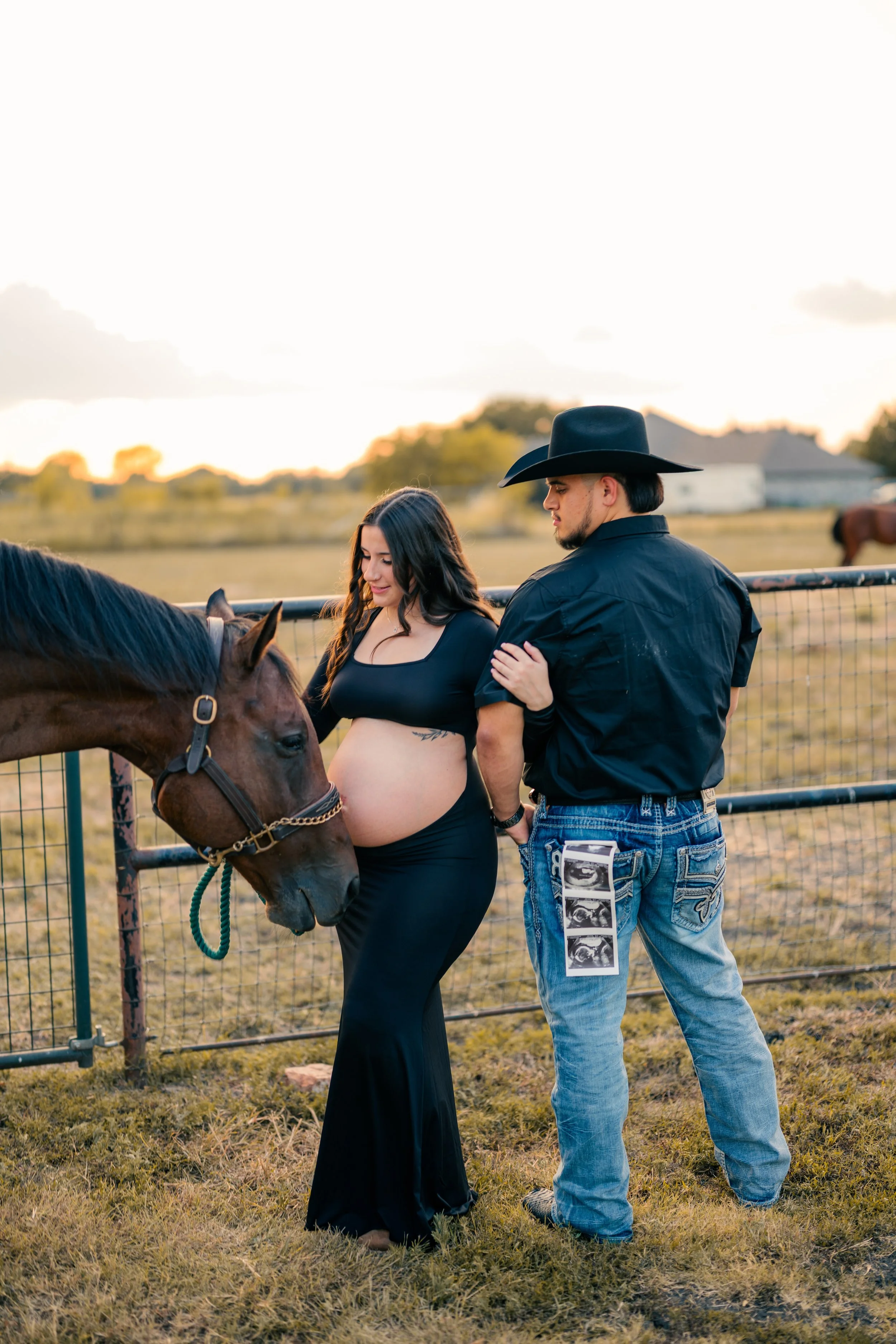 Pregnant woman in black dress with her belly exposed, standing next to a brown horse, with a man in cowboy hat and jeans touching her shoulder, in a rural outdoor setting during sunset.