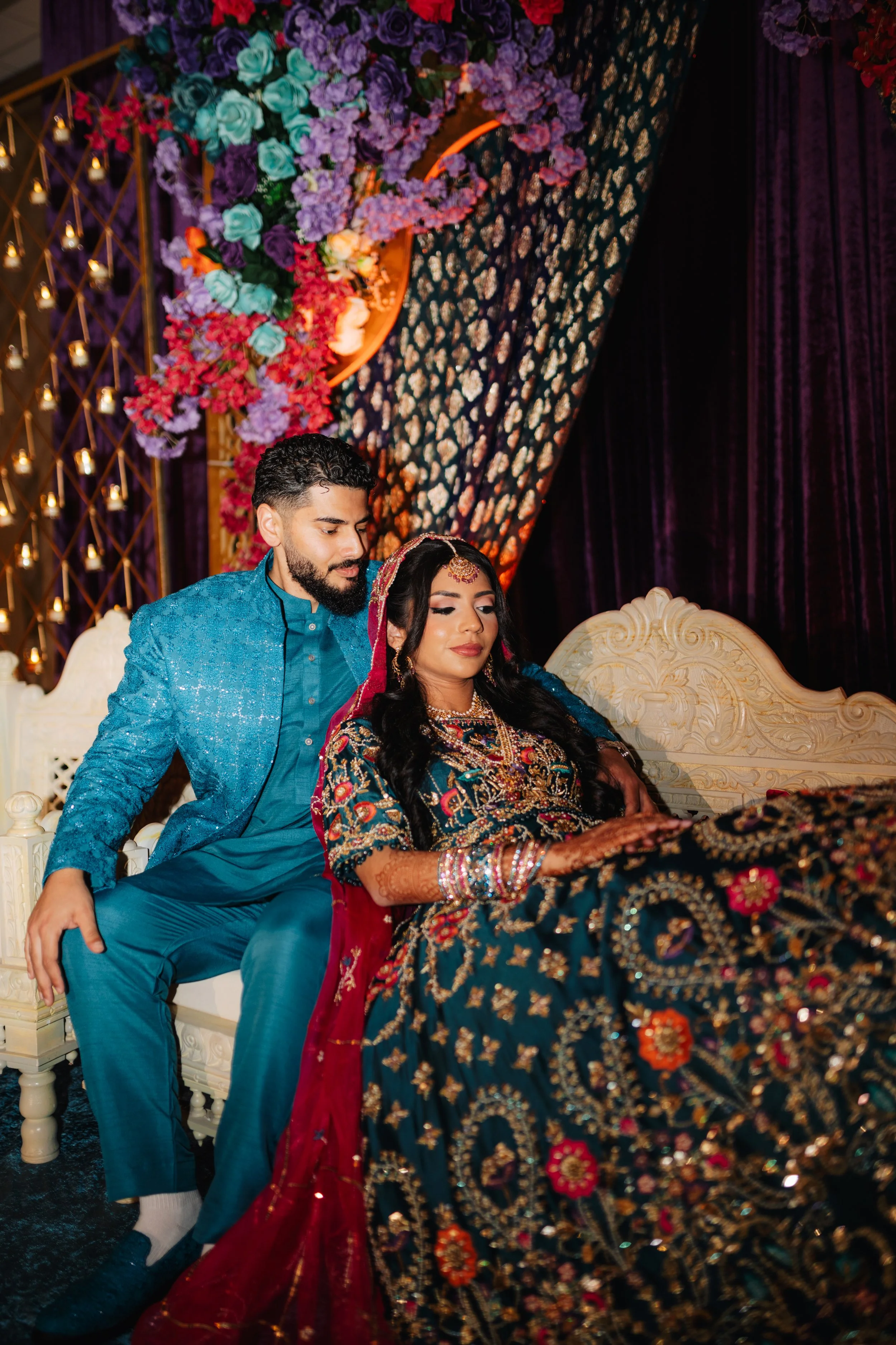 A couple in traditional Indian attire sitting on a decorative cream-colored sofa at a celebration event, with floral and curtain decorations in the background.
