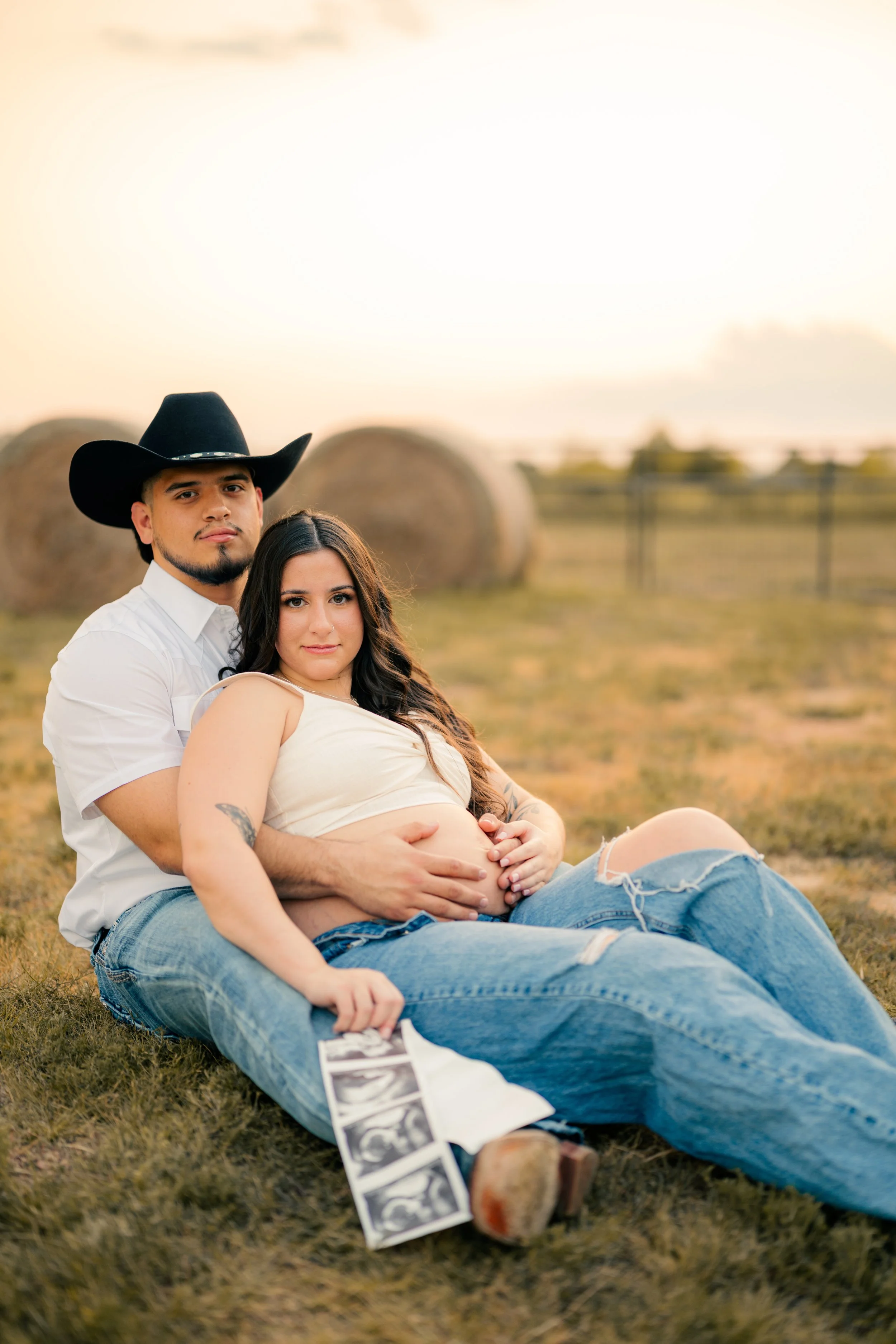 A pregnant woman and a man sitting on the grass in a rural field with hay bales in the background during sunset, with a strip of ultrasound images on the ground.