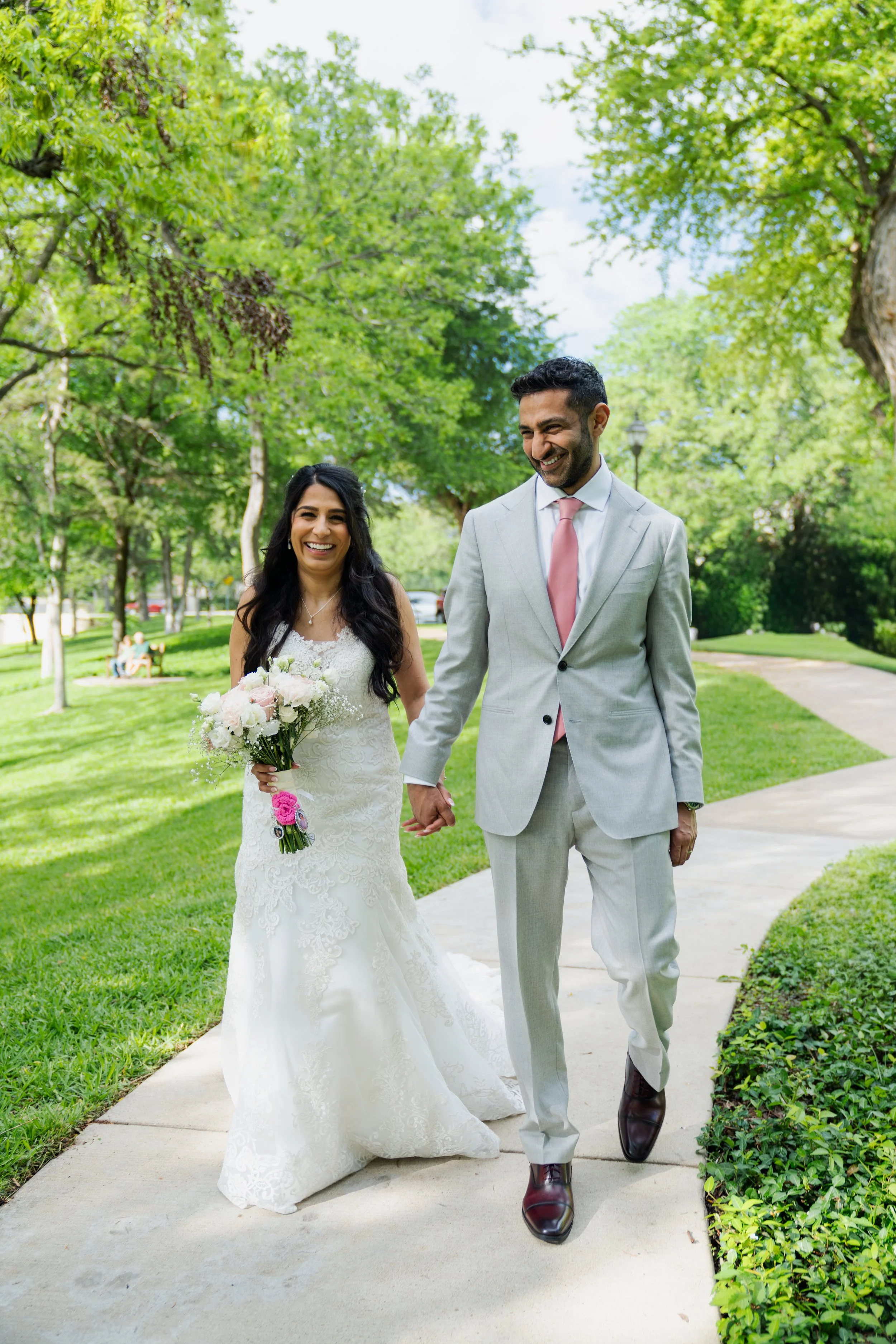 A newlywed couple, a woman in a white wedding gown holding a bouquet of flowers and a man in a light gray suit with a pink tie, walking hand in hand on a sidewalk in a park with green trees and grass, smiling and smiling.