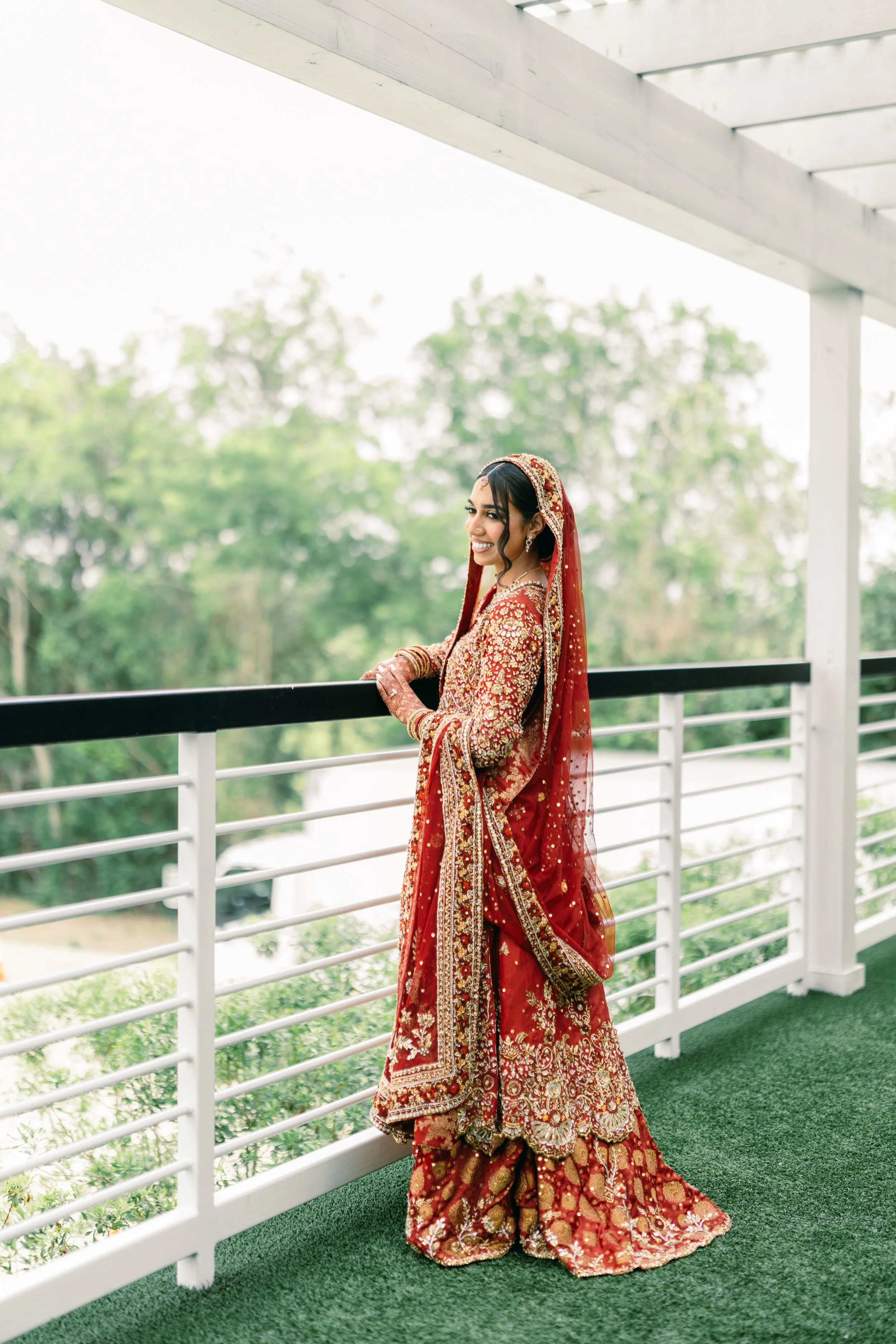 Woman in a red embroidered traditional Indian bridal dress with matching dupatta and jewelry, standing on a balcony with a white railing and green artificial grass, with greenery in the background.
