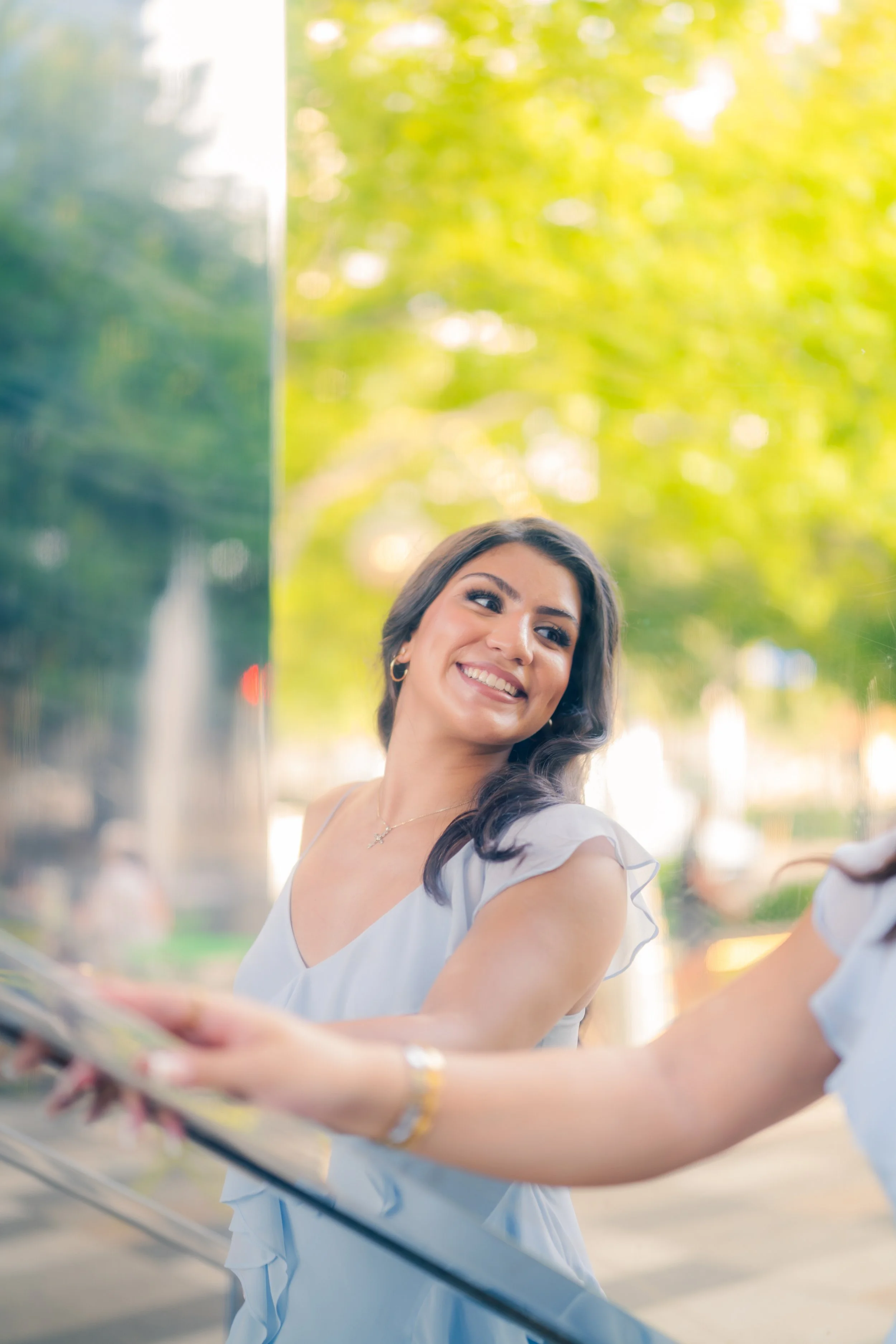 A smiling young woman with dark hair and earrings looks at her companion while standing near a glass barrier outdoors on a sunny day with green trees in the background.