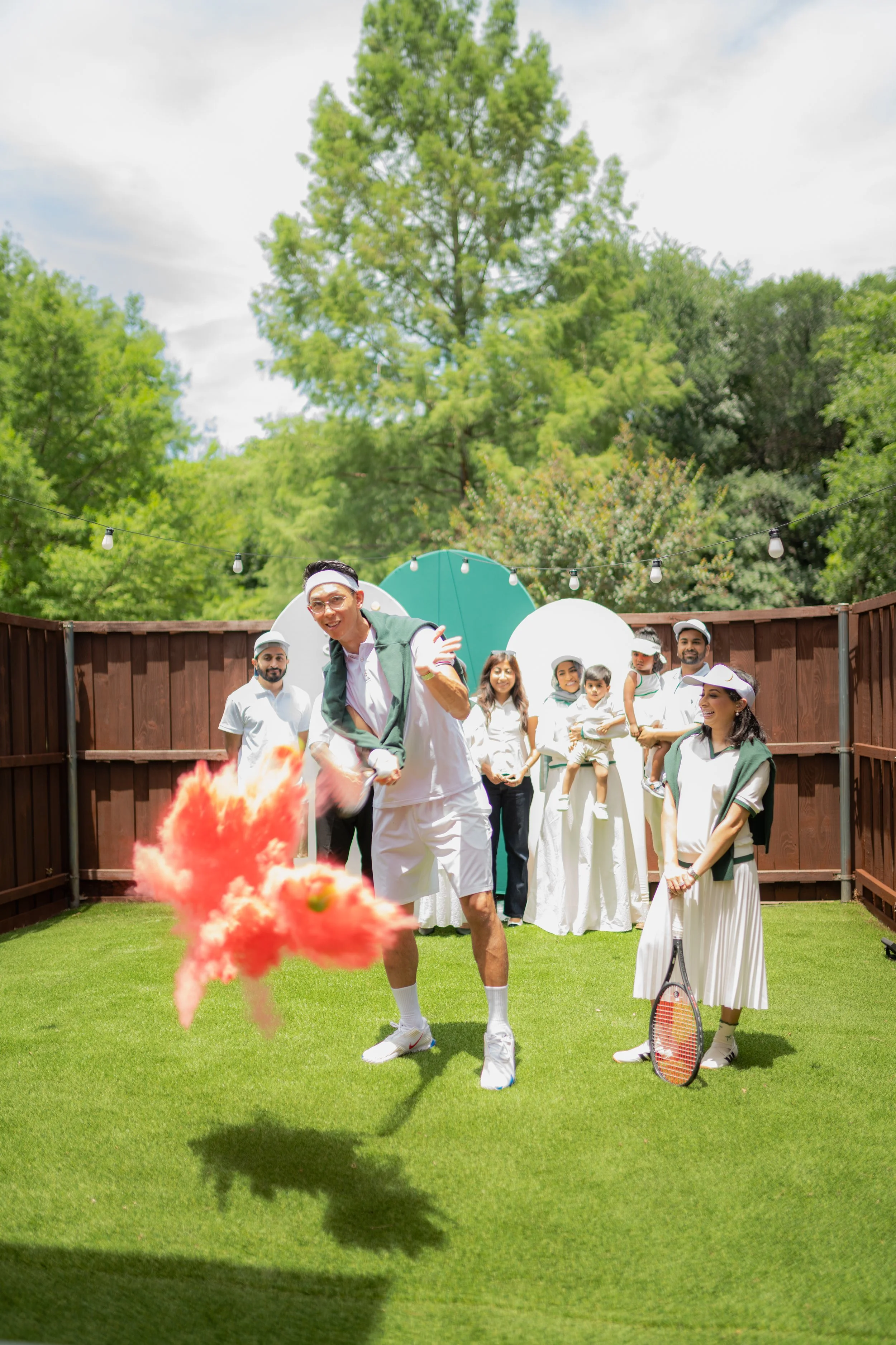 A group of people gathering in a backyard on a sunny day, some wearing athletic clothing, with a tennis racket visible, and a large tree in the background.