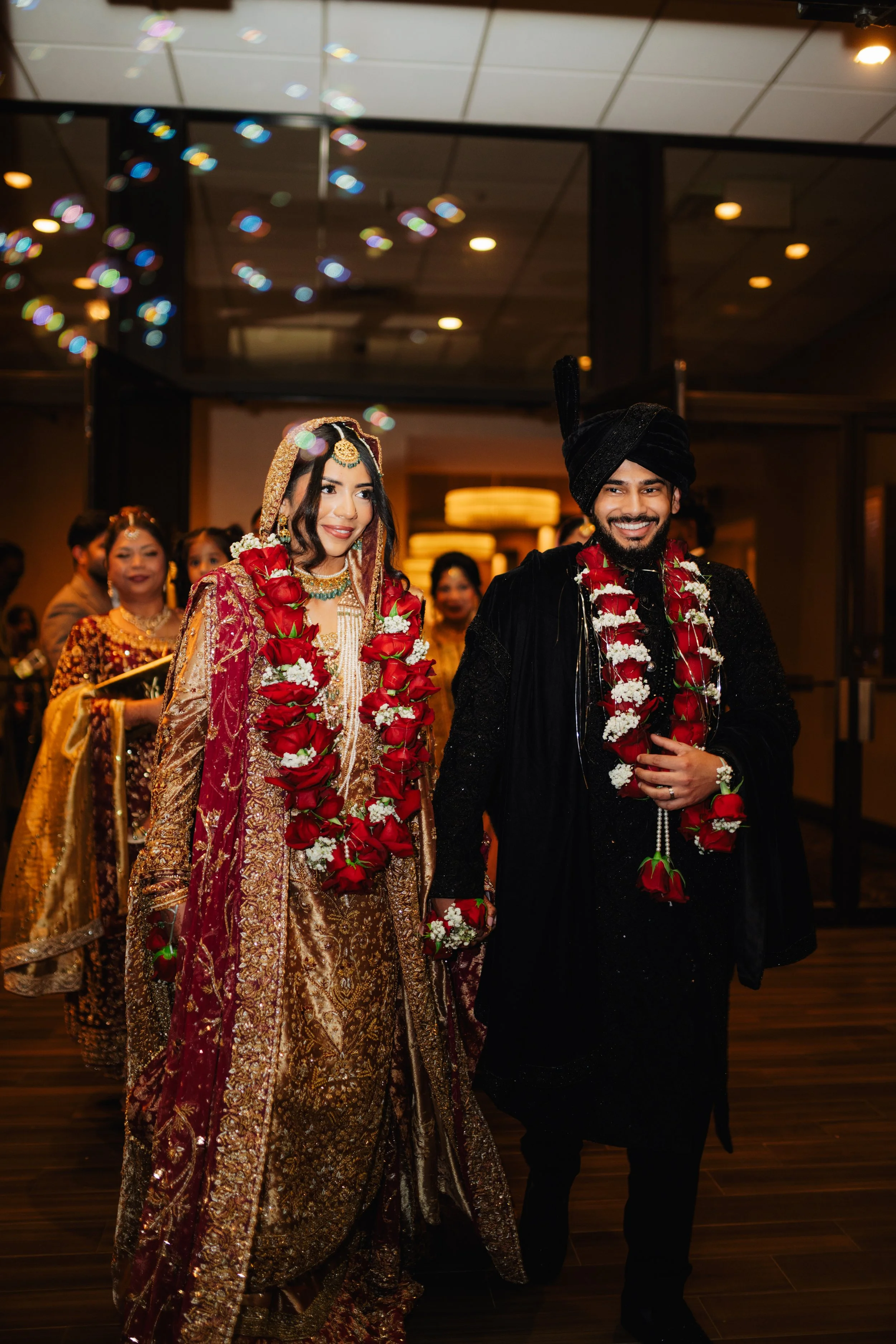 A bride and groom in traditional South Asian attire, wearing floral garlands, smiling at a wedding reception.