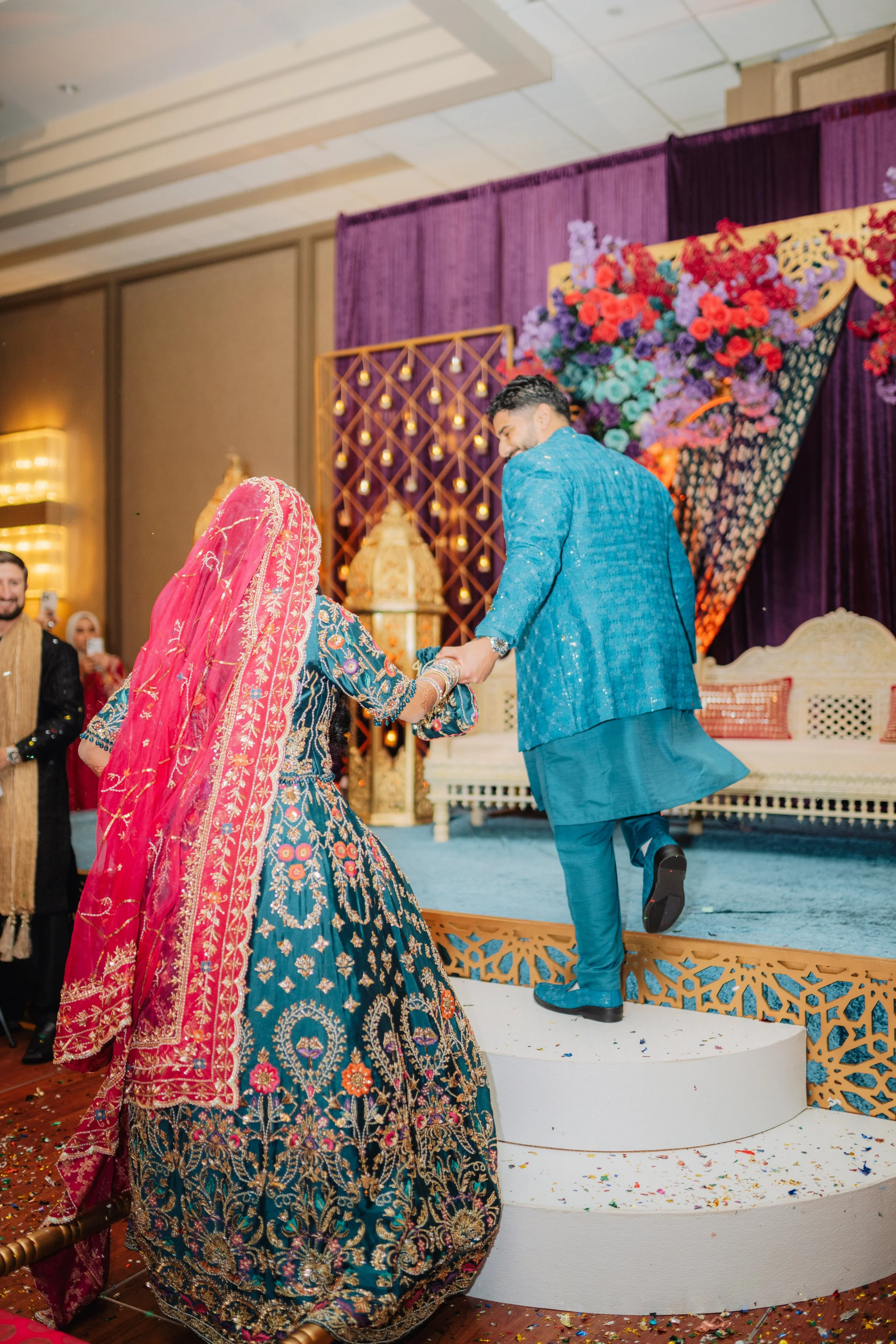 A groom in a blue traditional outfit is kneeling and holding hands with a bride dressed in a colorful embroidered wedding dress and a pink veil. They are on a decorated stage with purple drapes and a large floral arrangement in the background. Guests