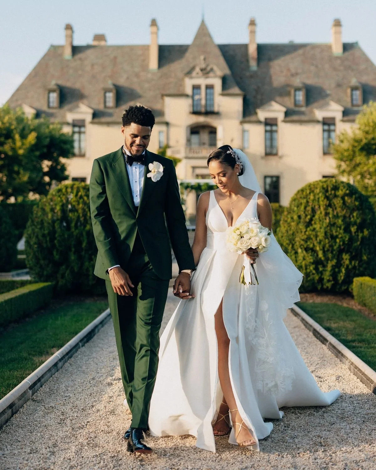 Bride in white gown holding bouquet and groom in green suit walking hand-in-hand in front of a large estate.