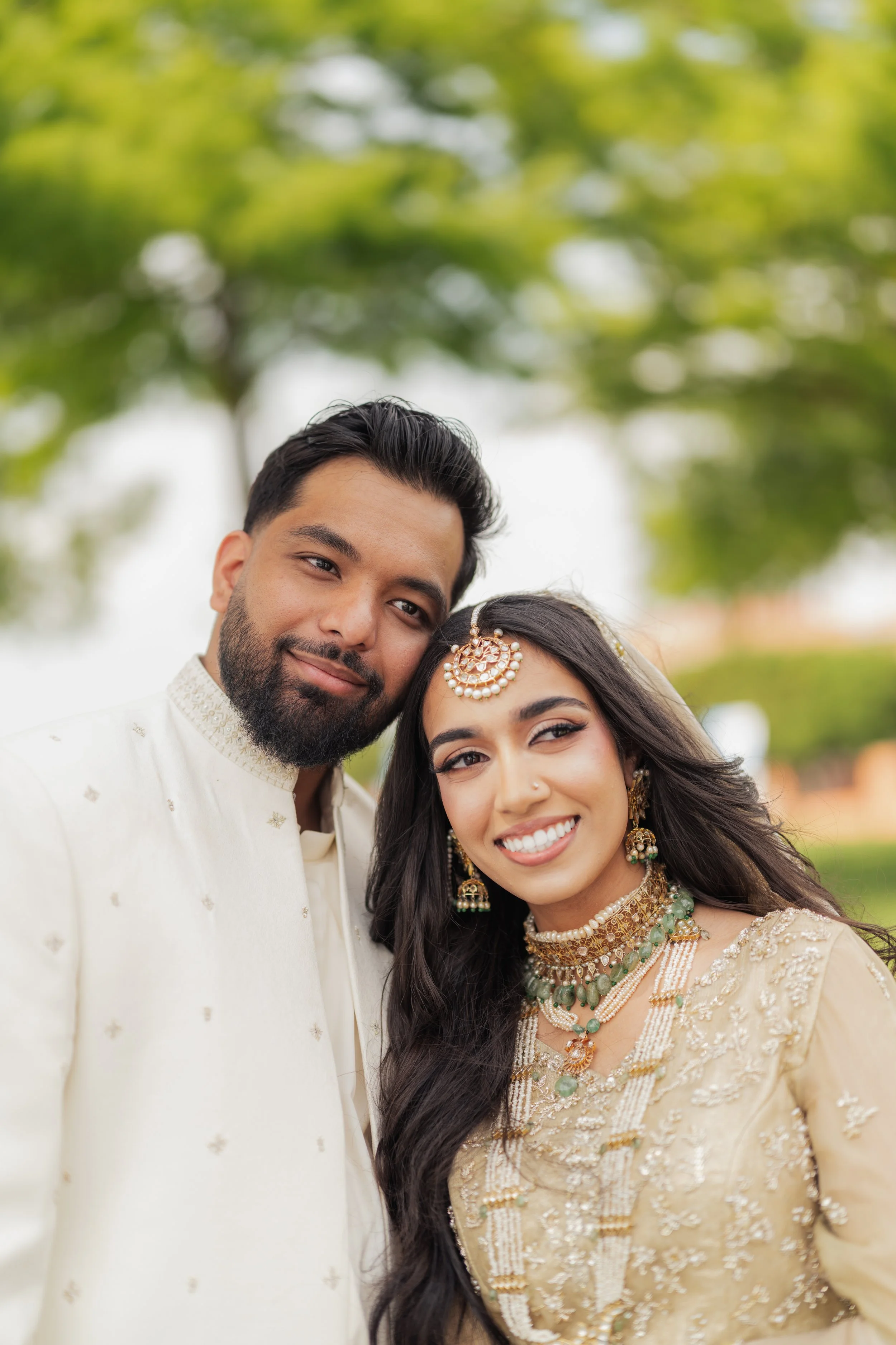 Close-up of a joyful couple in traditional Indian attire, outdoors with blurred green trees in the background.