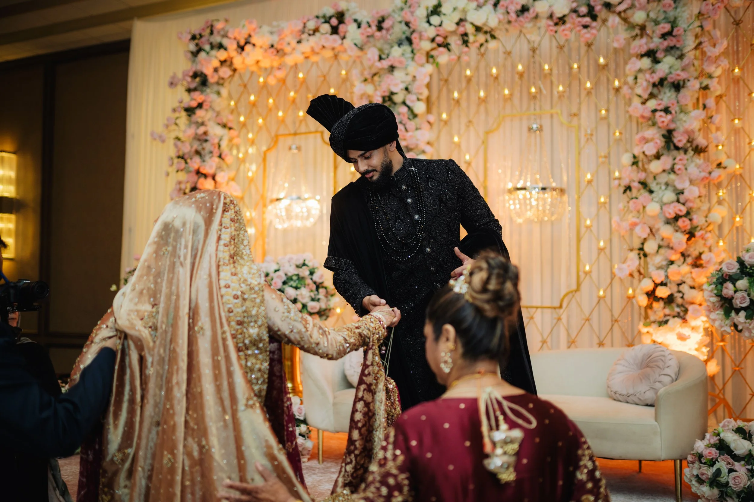Bride and groom during a traditional Indian wedding ceremony, with the groom wearing a black sherwani and turban, and the bride dressed in a gold and pink embroidered saree, surrounded by guests, with a floral backdrop and warm lighting.