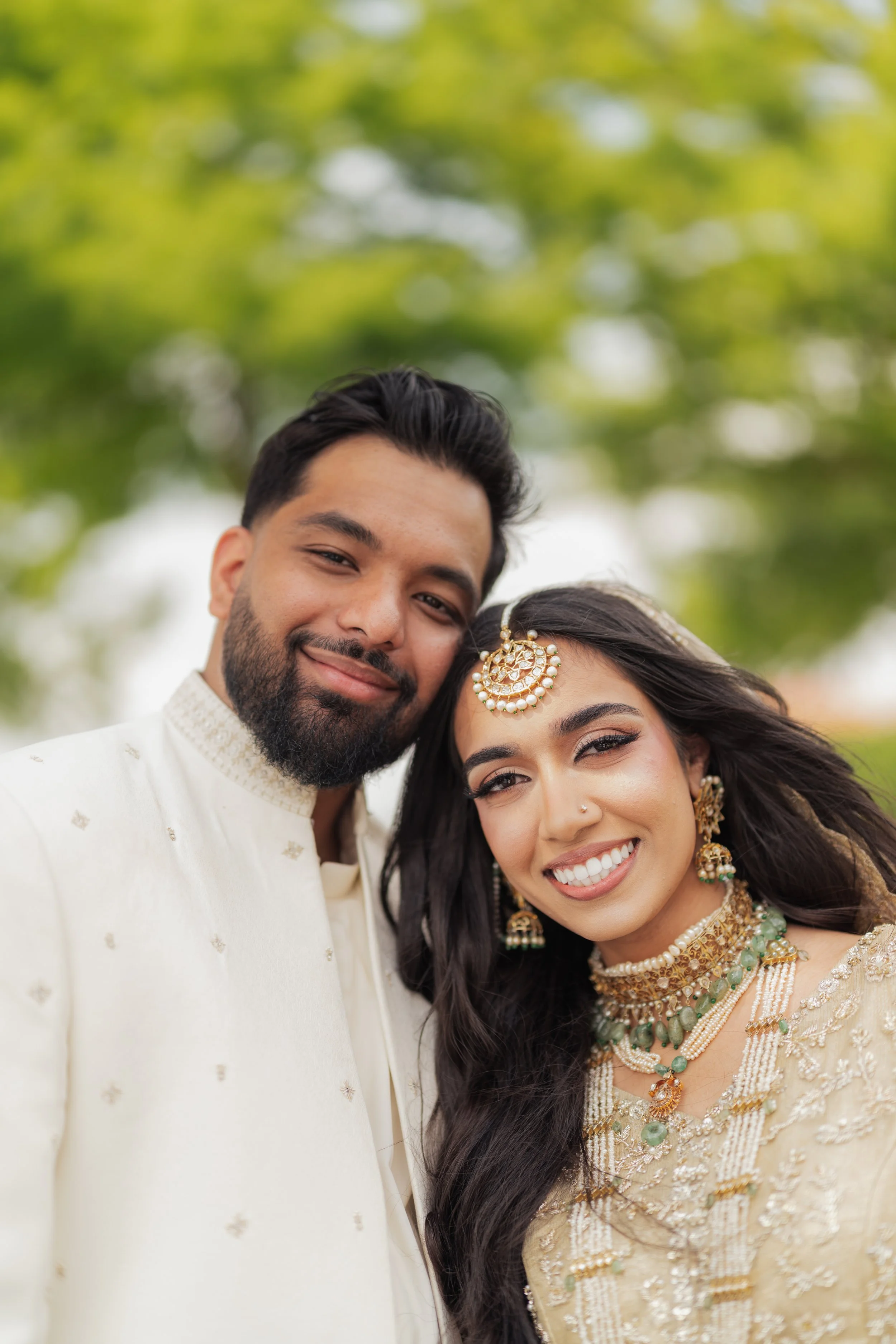 A happy couple dressed in traditional Indian wedding attire smiling outdoors with green trees in the background.