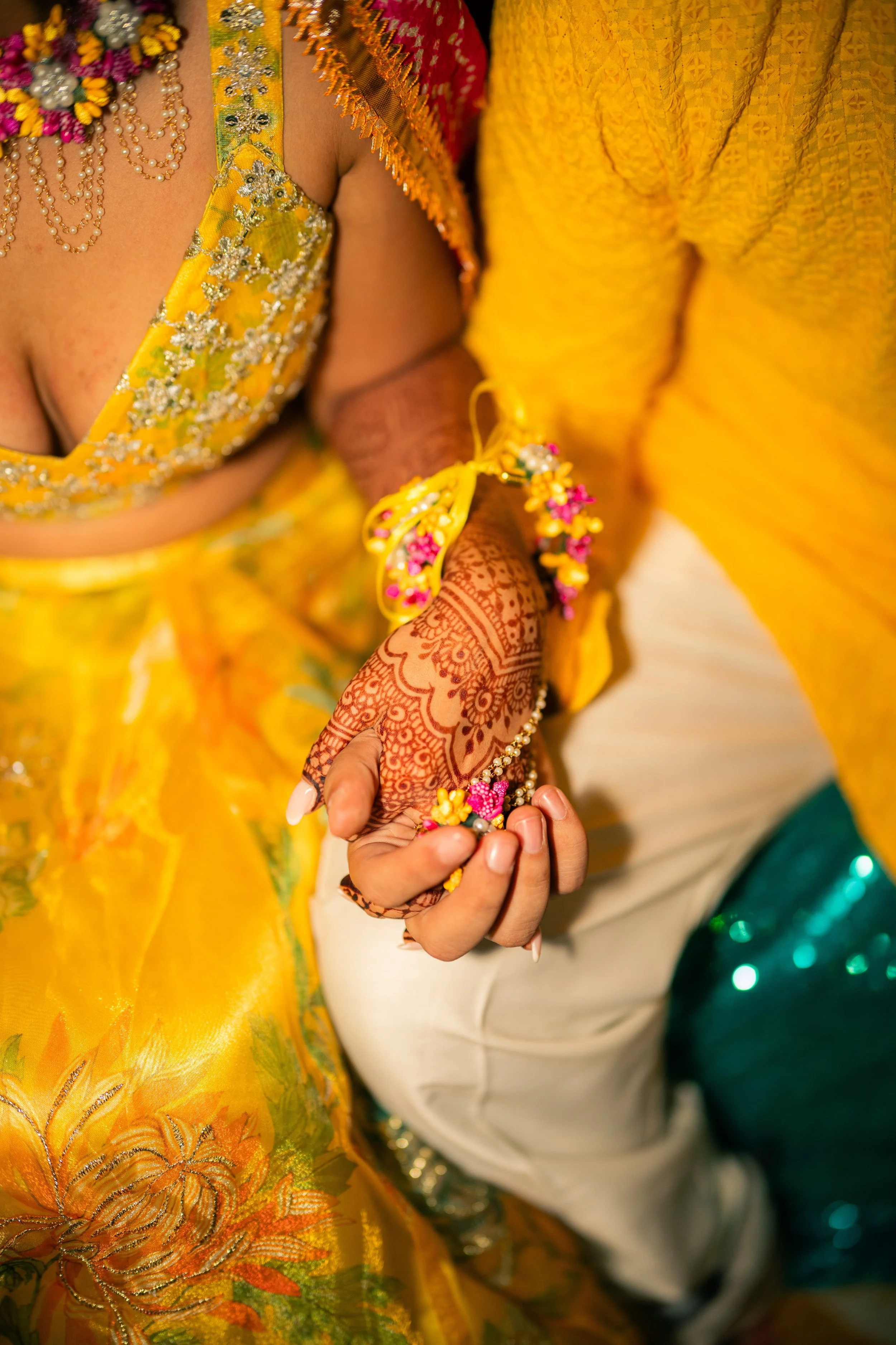Close-up of a couple holding hands, with the woman displaying henna and wearing traditional jewelry, in colorful clothing at a wedding or celebration.