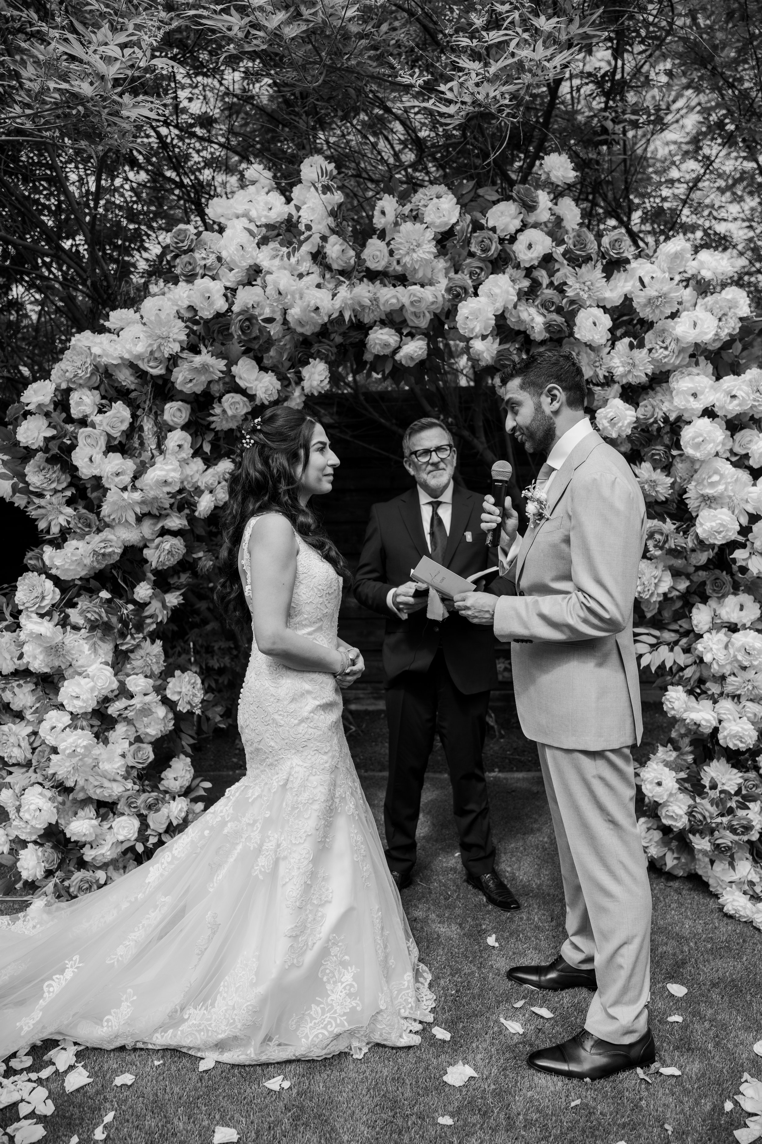 A bride and groom standing face to face during a wedding ceremony outdoors, with a floral arch and a man officiant in the background.