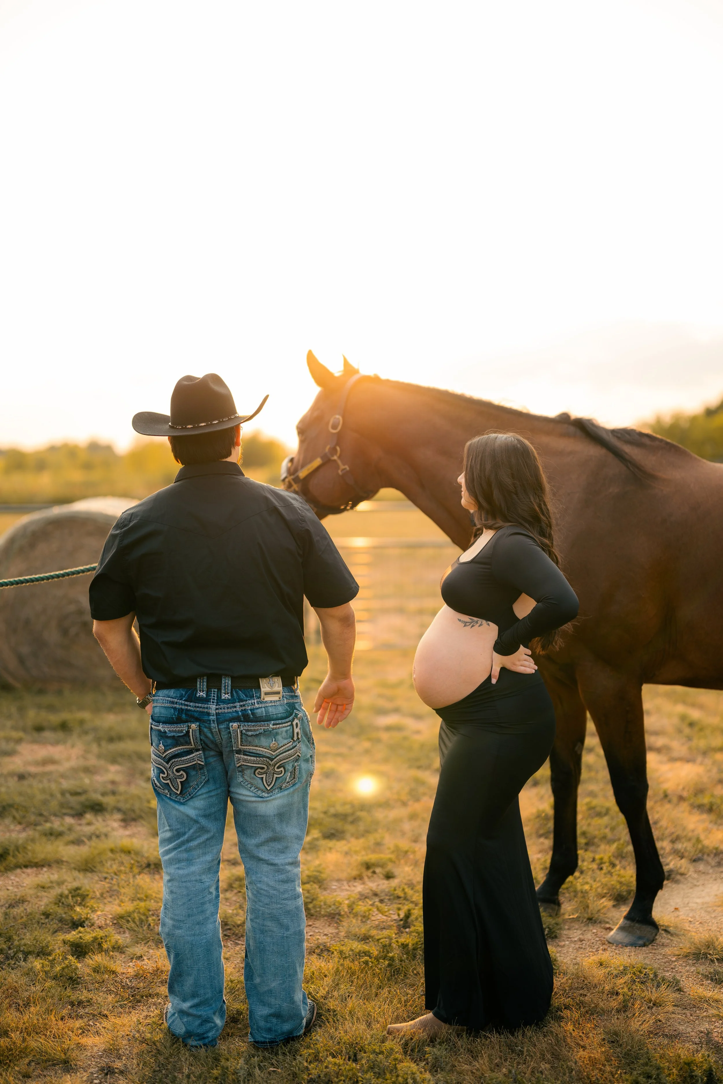 A pregnant woman and a man standing next to a horse outdoors during sunset.