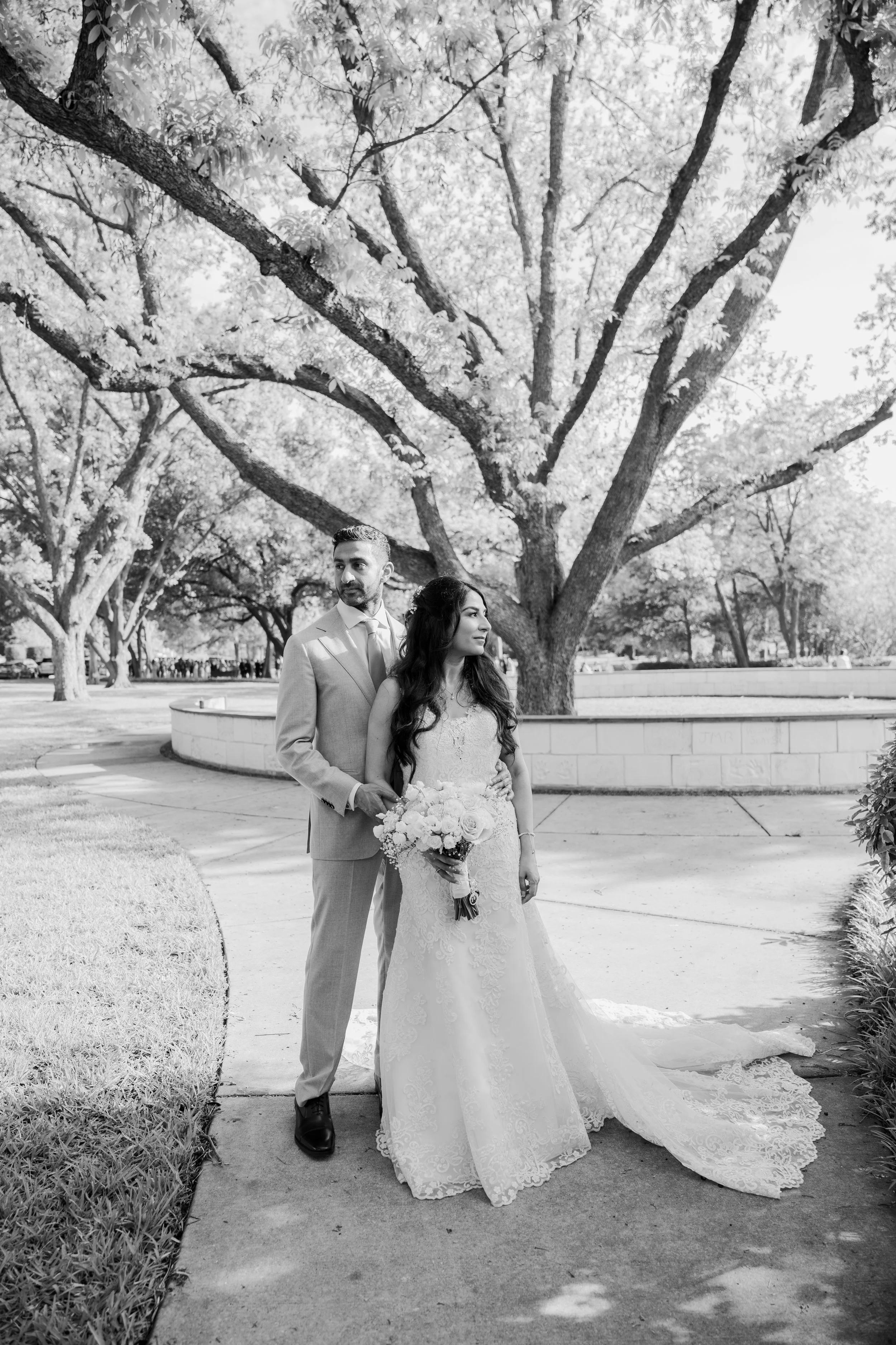 A bride and groom in wedding attire standing together on a sidewalk in a park with tall trees and people in the background. The bride is holding a bouquet of flowers, and both are looking away from the camera.