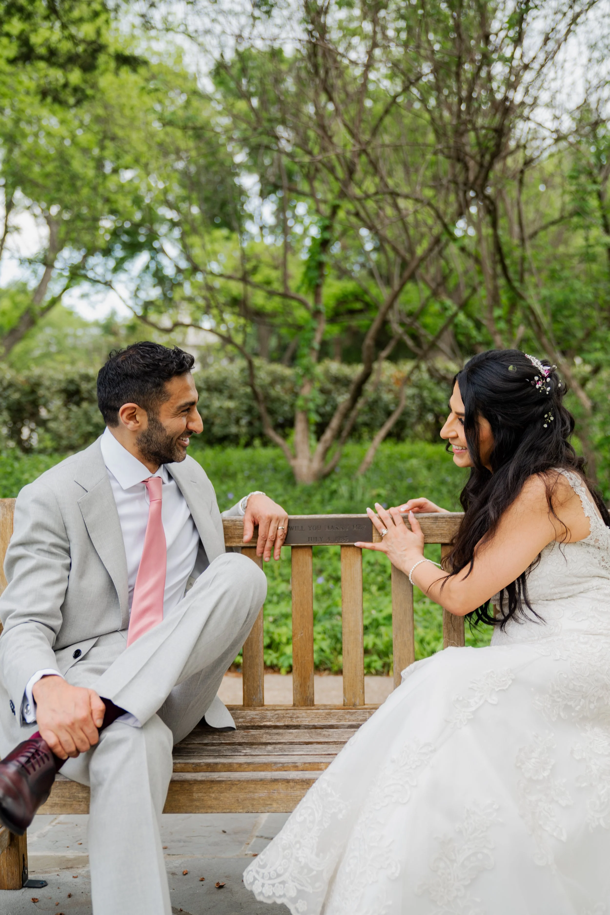 A man and woman sitting on a park bench, the man wearing a light gray suit with a pink tie, and the woman in a white wedding dress with long, dark hair and floral hair accessories, smiling at each other outdoors surrounded by trees.