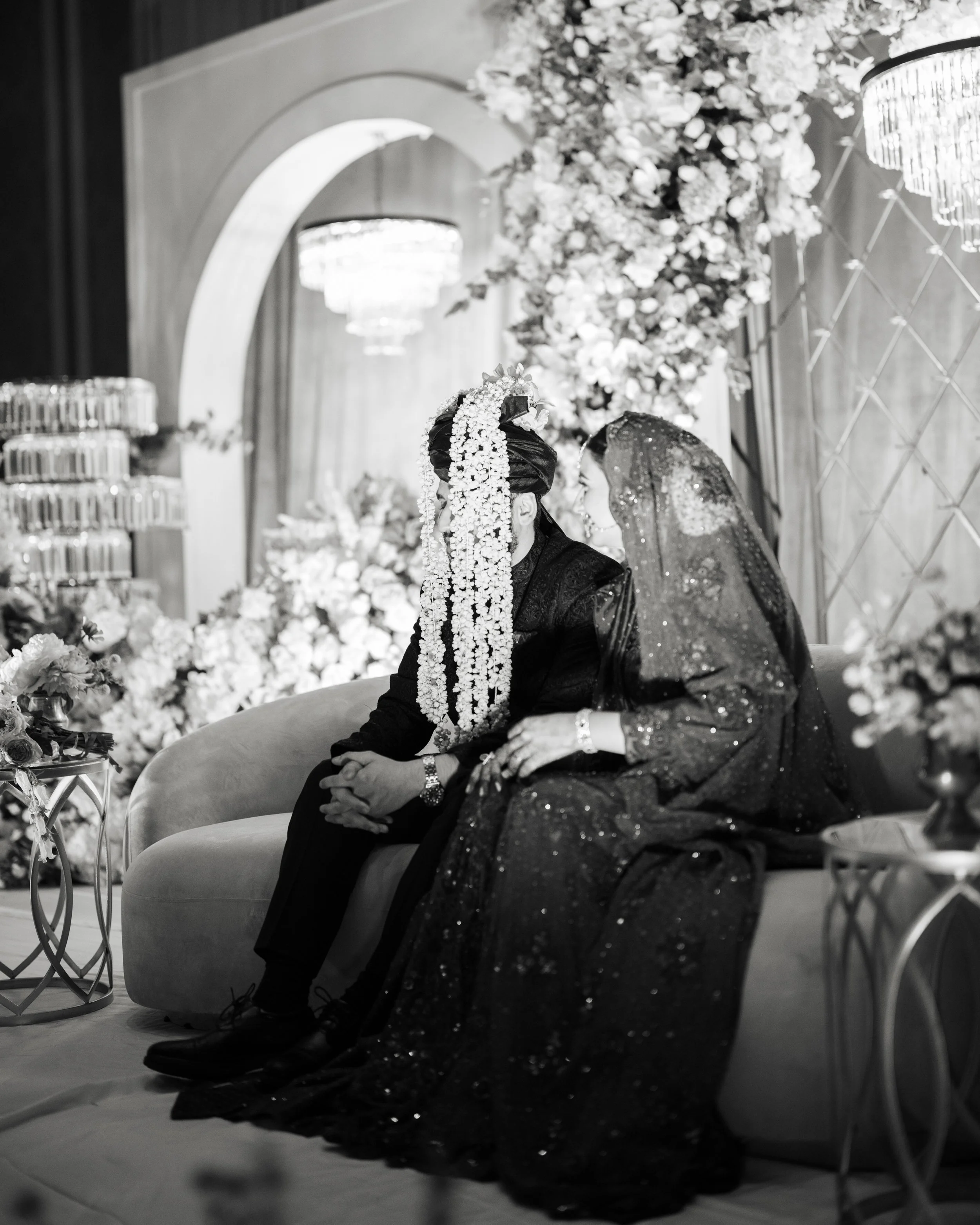 Two women in traditional attire sitting on a sofa during a wedding ceremony, decorated with flowers and chandeliers.