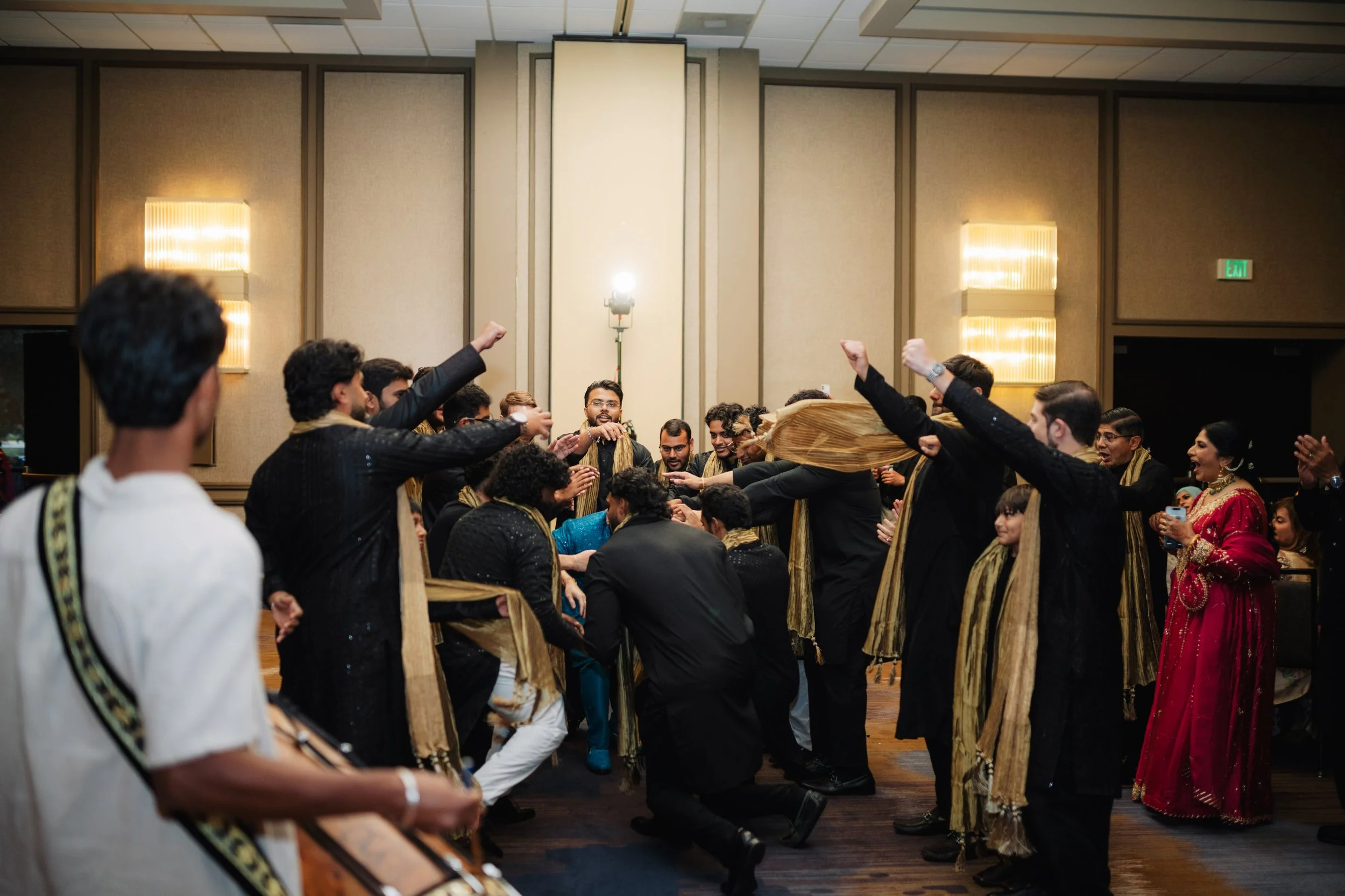 Group of people dressed in traditional South Asian attire dancing and celebrating in a banquet hall.